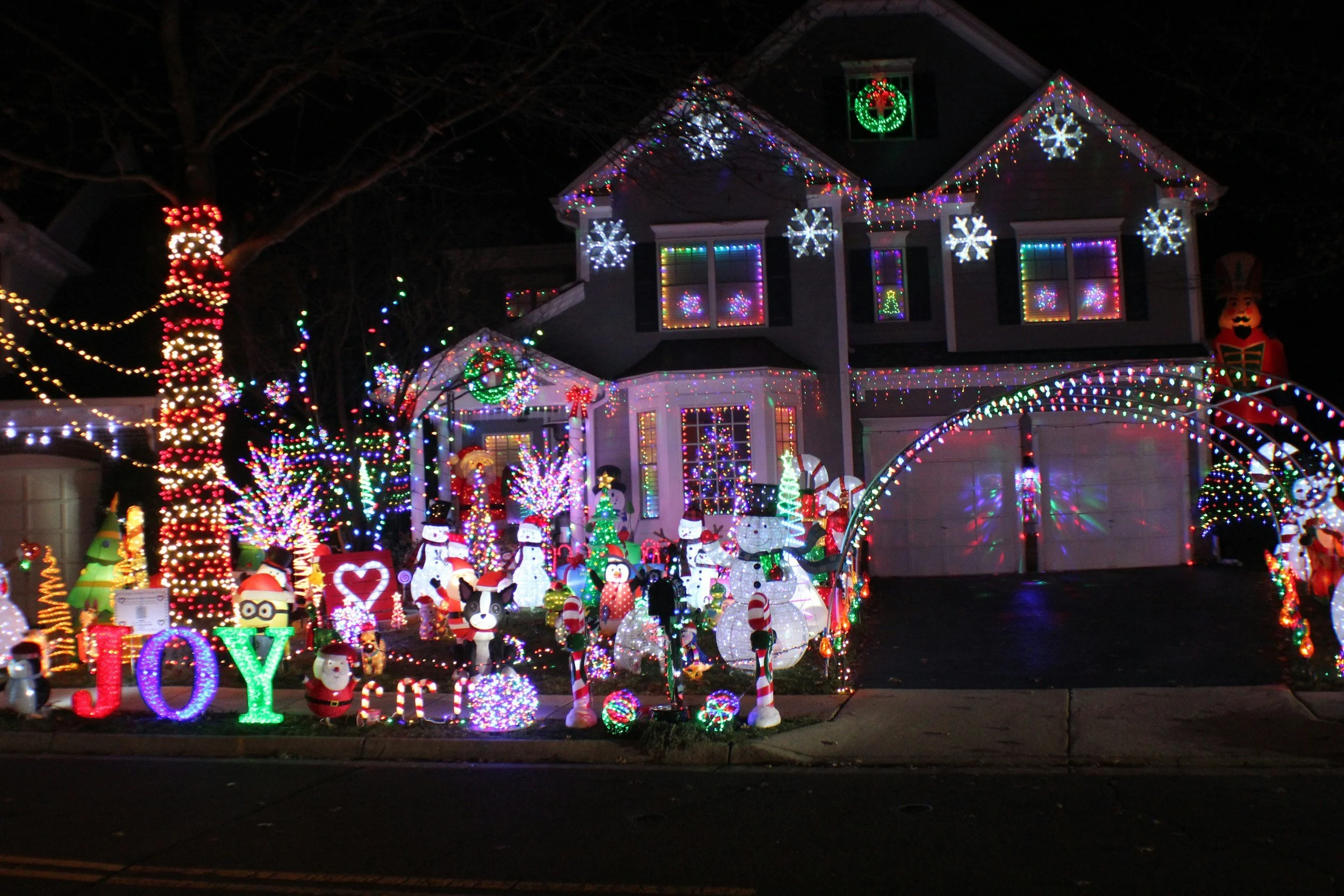 A house decorated with colorful Christmas lights and inflatable holiday figures, including snowmen, Santa Claus, and a penguin, with a yard sign spelling 'JOY'.