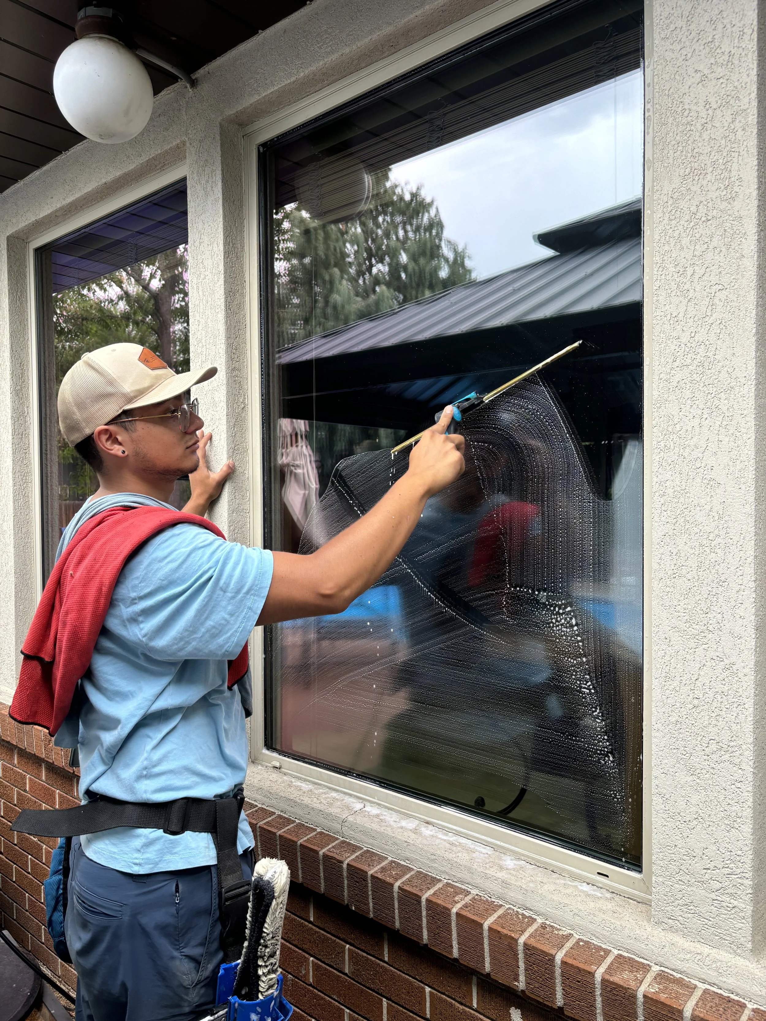 A person cleaning a large window with a squeegee, wearing a baseball cap, glasses, a light blue shirt, and a red towel draped over his shoulder.