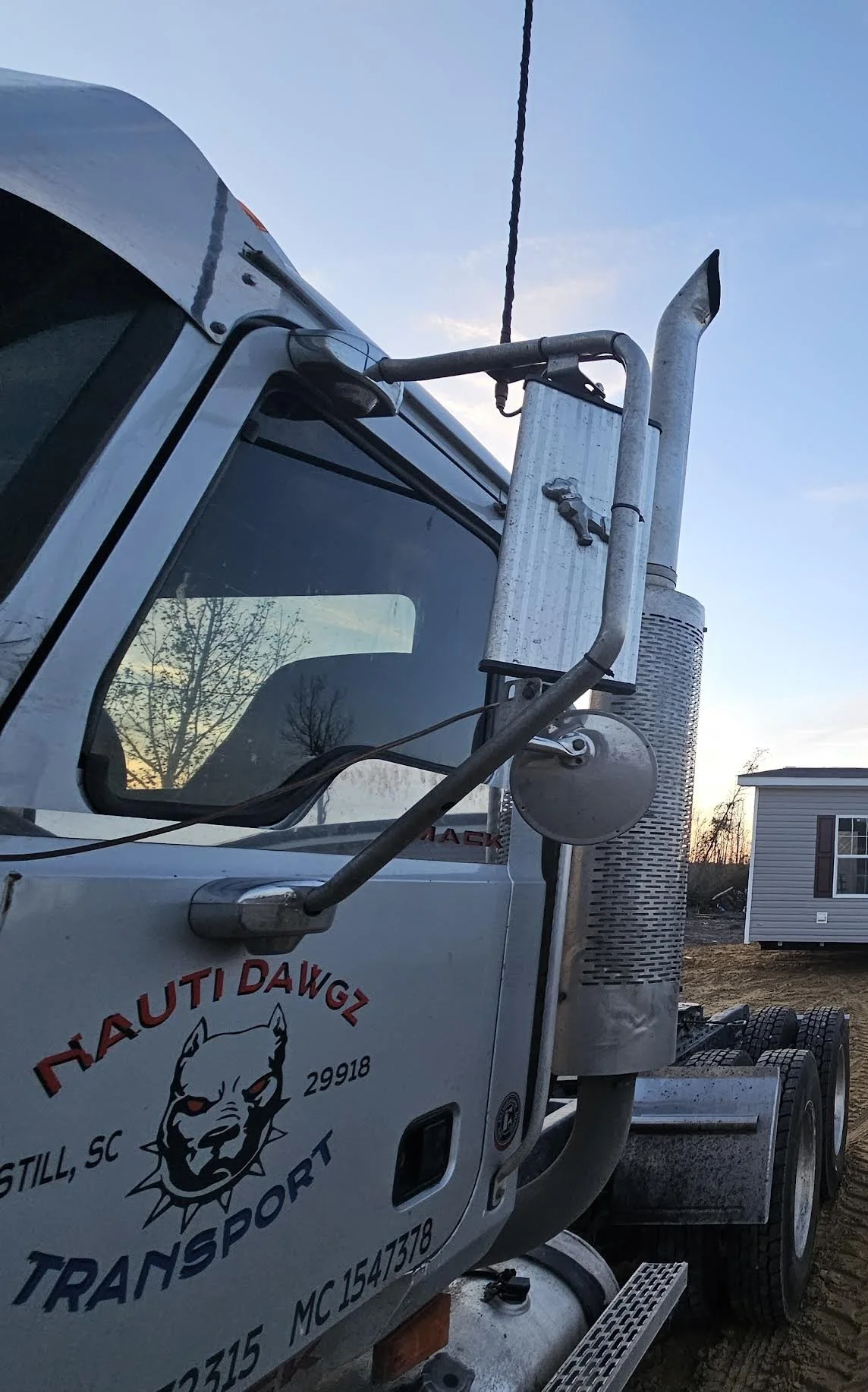 Close-up of the front side of a white truck with business logo and contact information, parked outdoors during sunset with a small house and bare trees in the background.