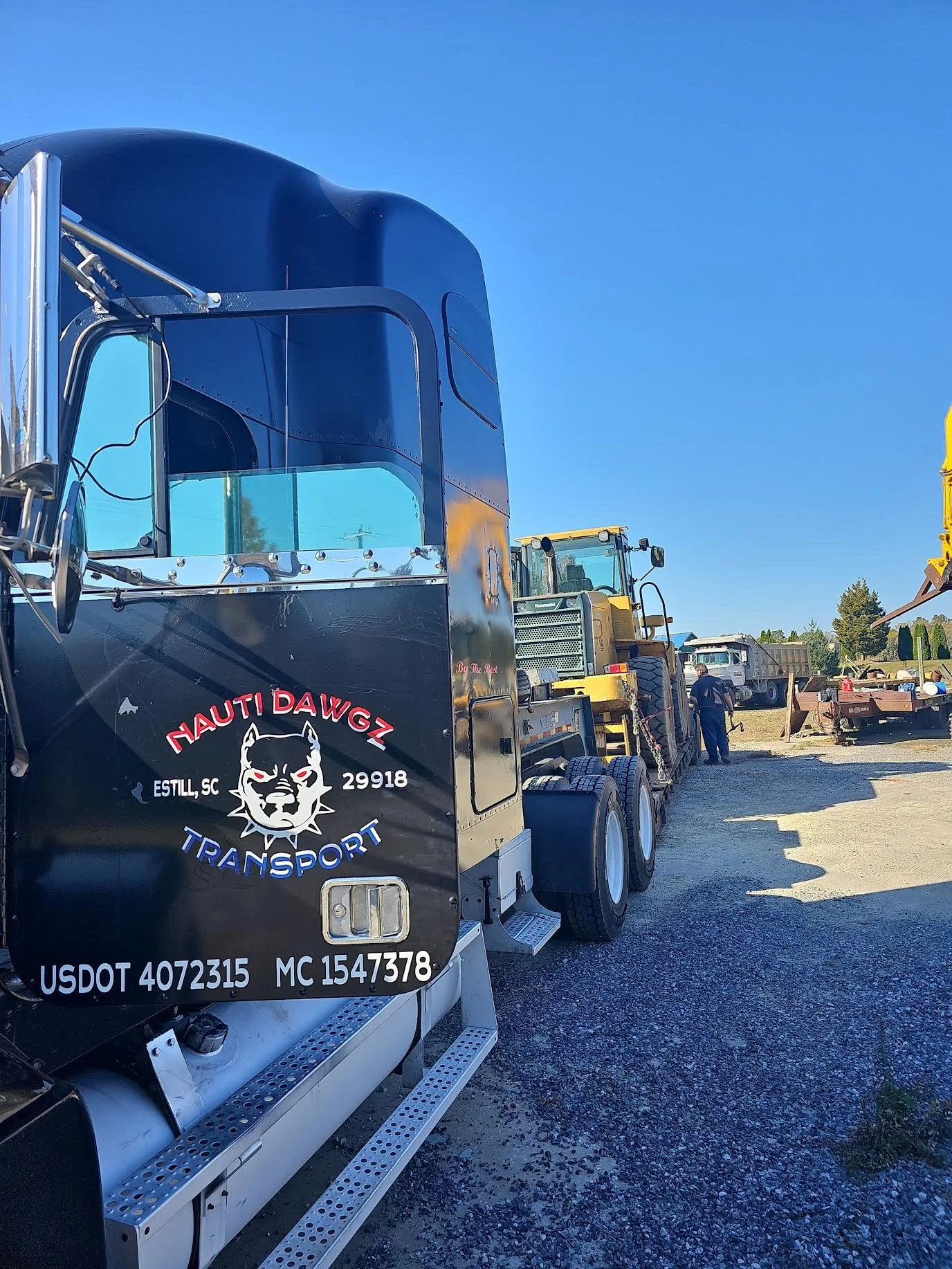 A large black semi-truck with a logo of a fierce dog face on the door, parked outdoors on a gravel lot under a clear blue sky. Several other trucks and workers are visible in the background.
