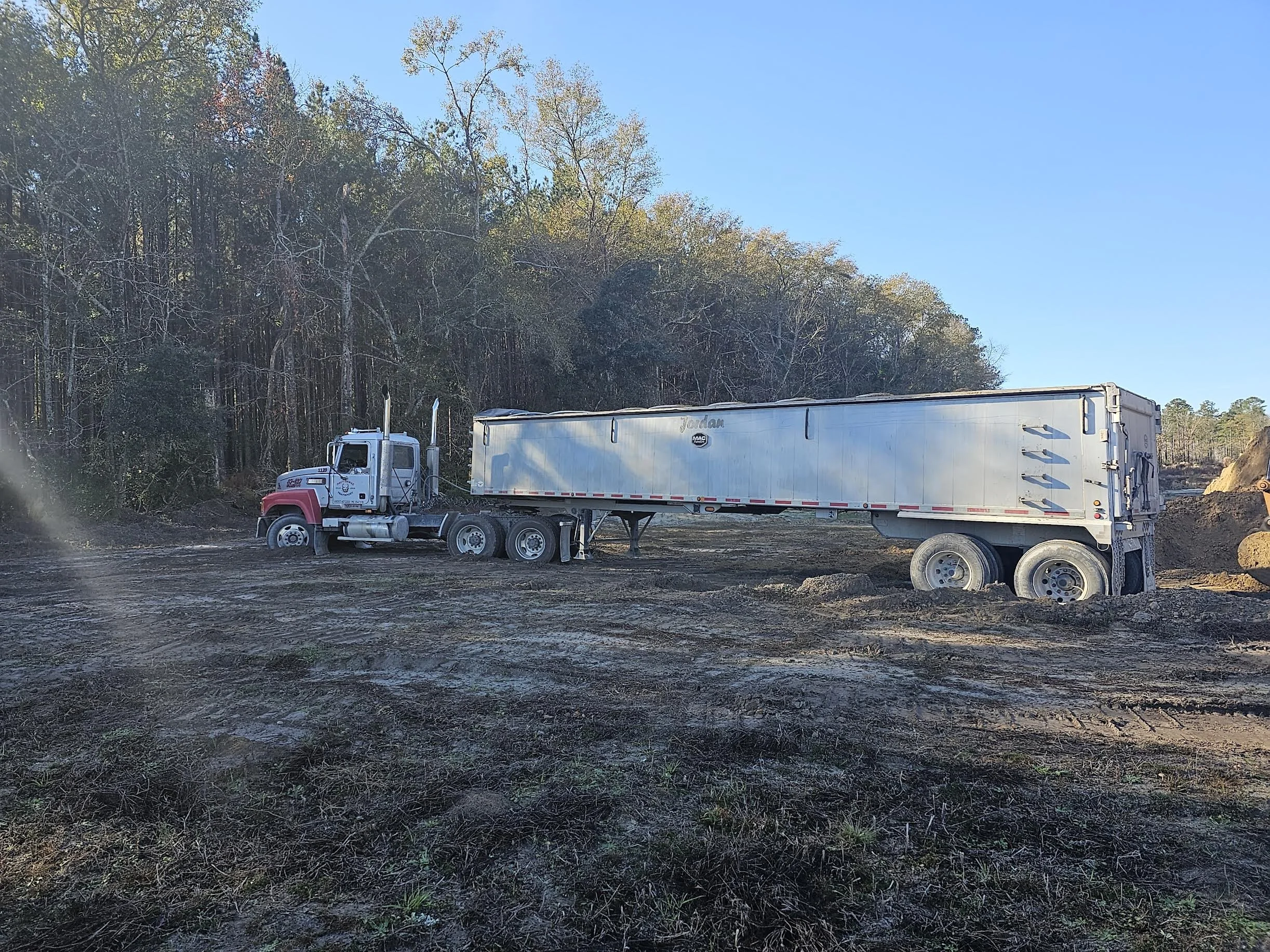 A large semi-truck with a red and white cab and a white trailer, parked on a dirt area near a wooded hillside under a clear blue sky.
