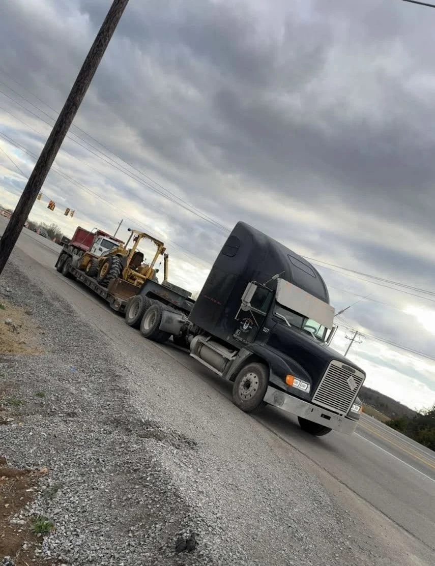 A black semi-truck parked on the side of a road, carrying a yellow bulldozer and a red truck bed on its flatbed trailer. Overcast sky with dark clouds.