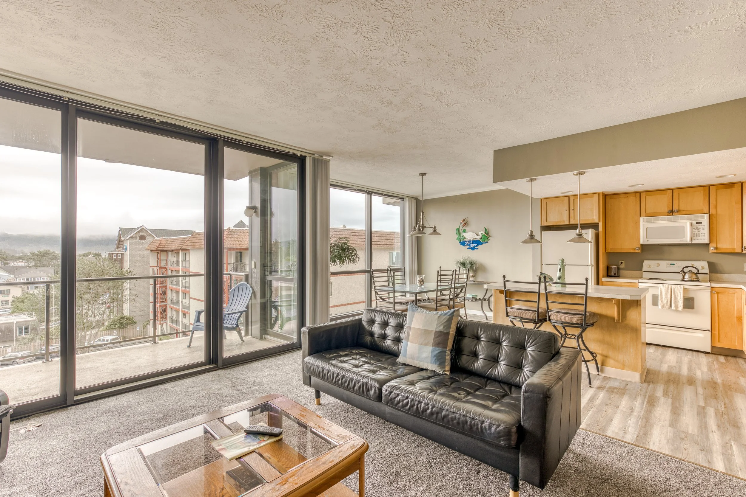 Modern living room with large windows, a black leather sofa, glass coffee table, open kitchen with light wood cabinets, dining table, and balcony view.