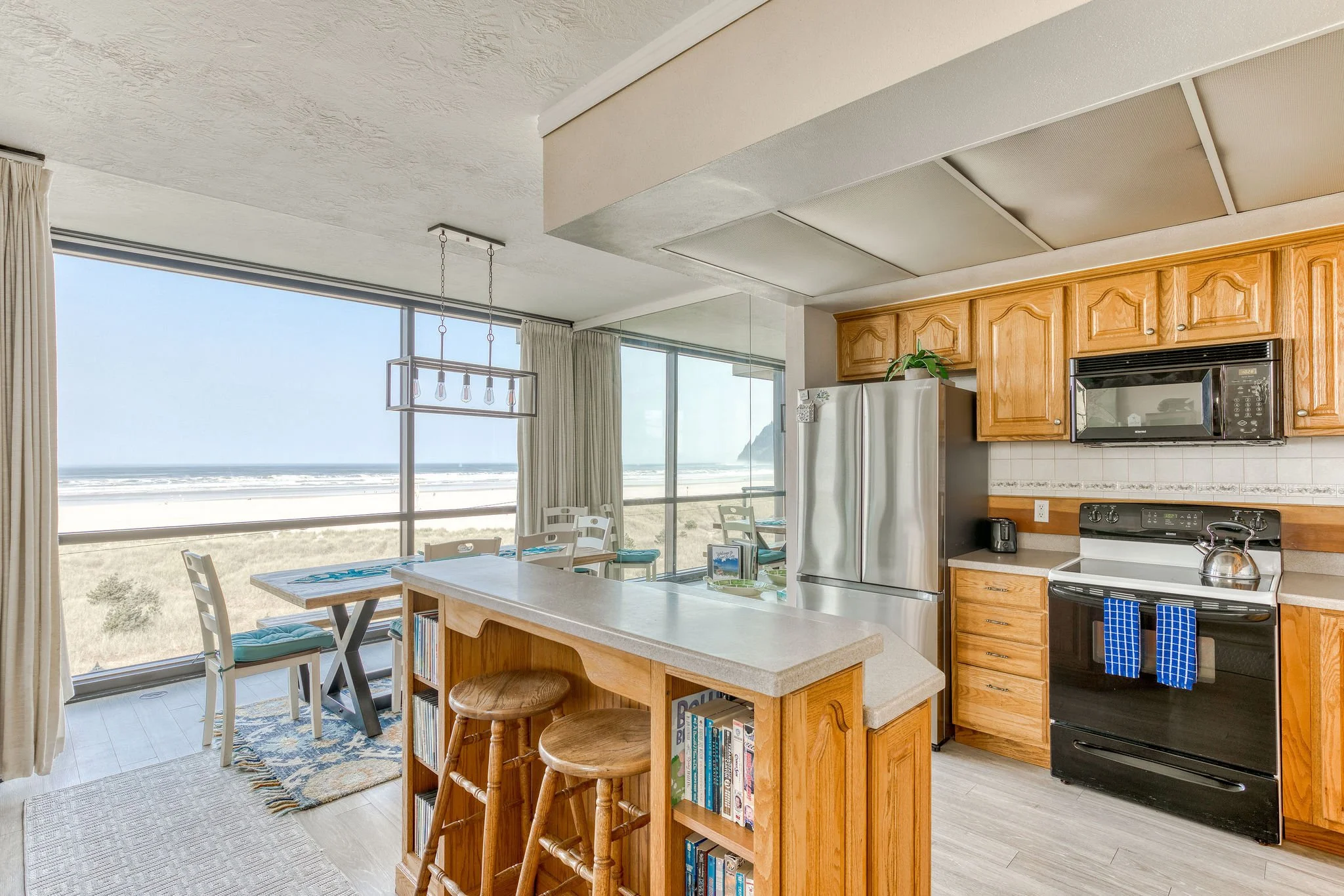 Beachfront kitchen and dining area with ocean view, wooden bar, stools, stainless steel appliances, and light wood cabinetry.