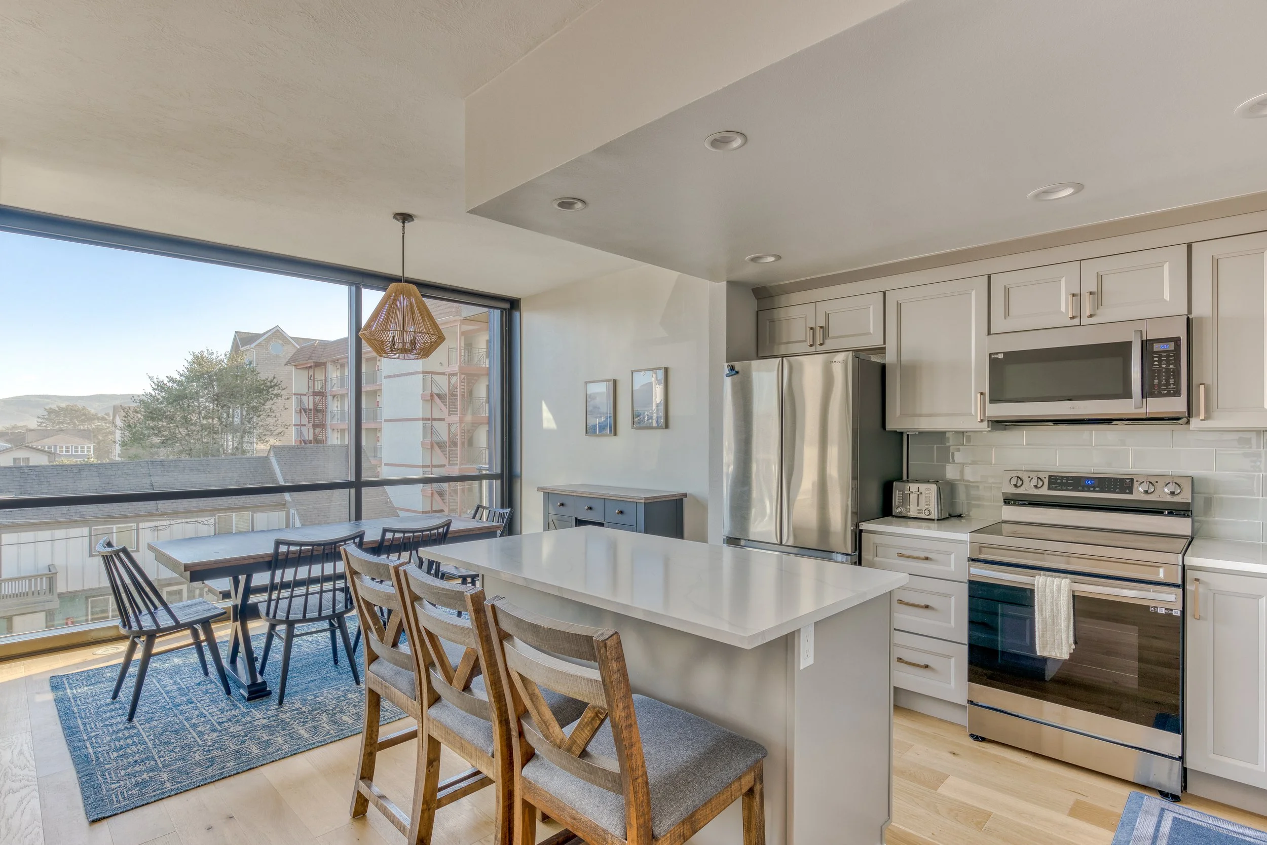 Modern kitchen and dining area with large windows, white cabinets, stainless steel appliances, and wooden chairs, featuring a pendant light and view of neighboring buildings.