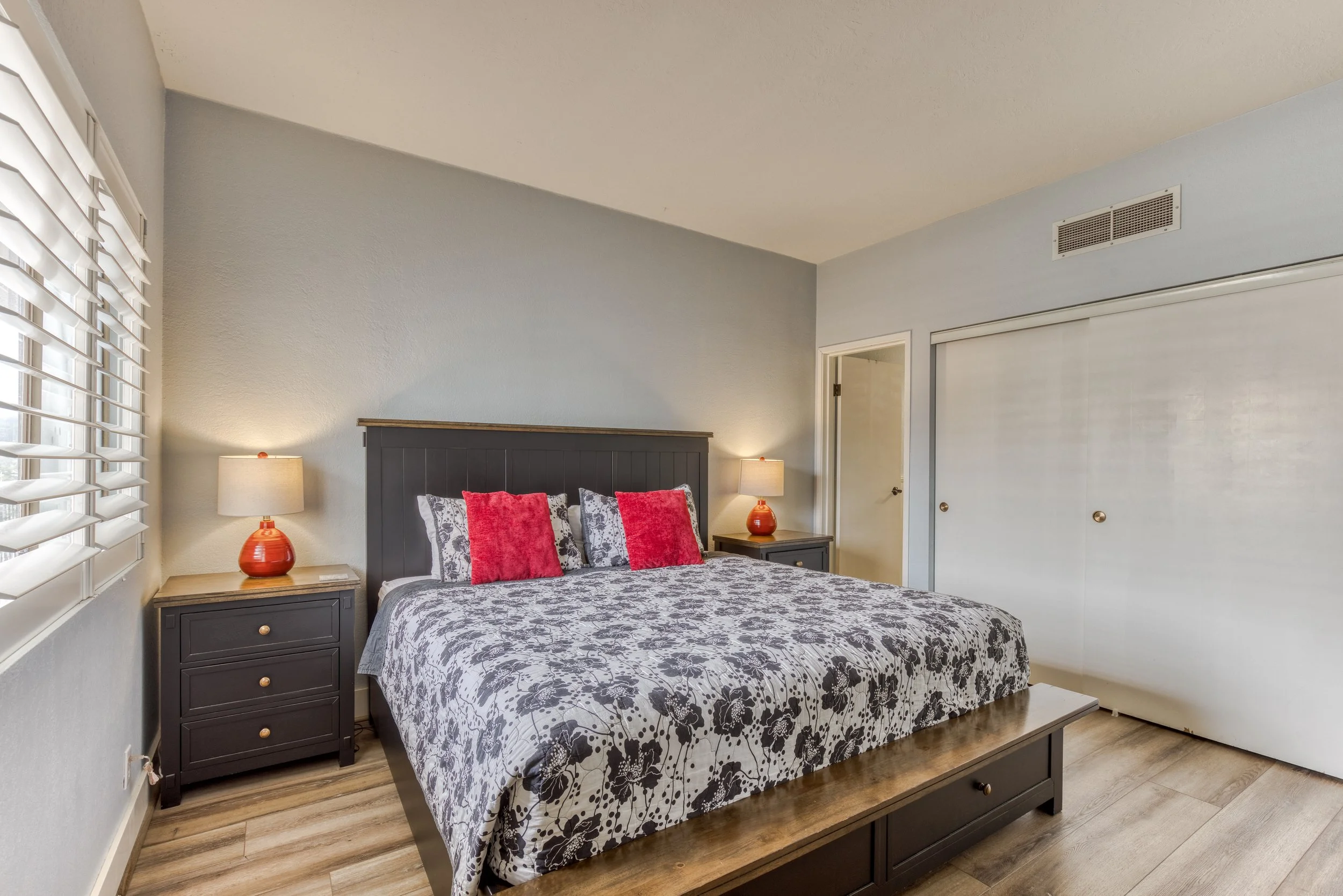 Modern bedroom with a large bed featuring a floral patterned bedspread, red accent pillows, and wooden nightstands. The room includes two lamps and a closet with sliding doors.