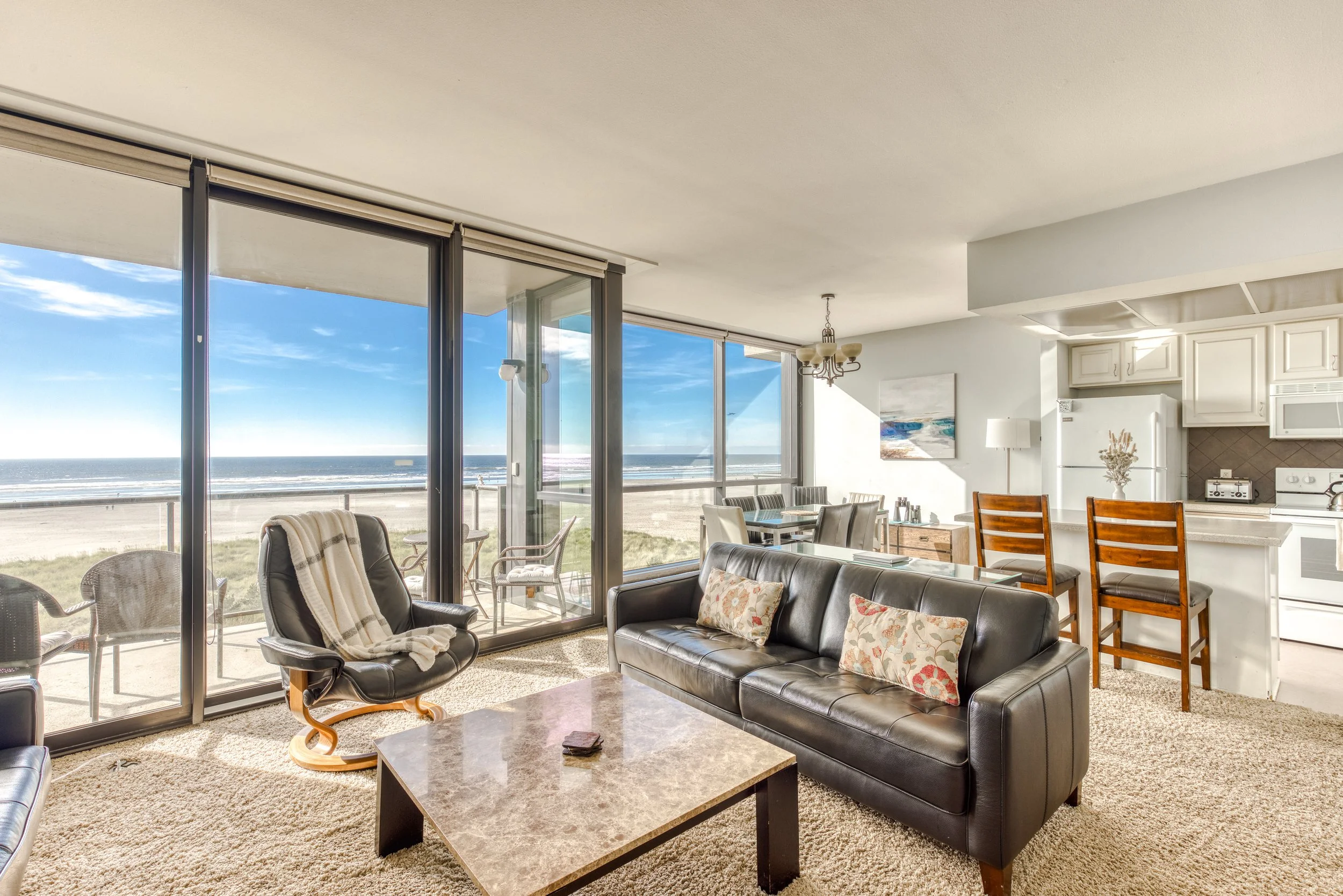 Coastal living room with ocean view, glass doors, leather sofa, dining area, and kitchenette.