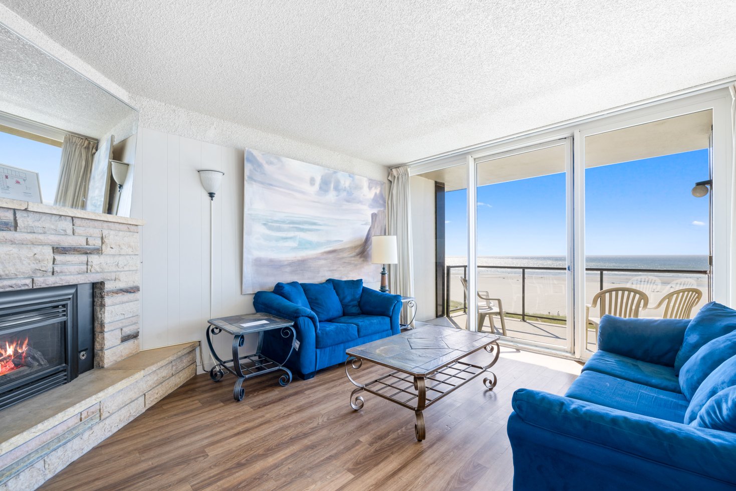 Coastal living room with blue sofas, large seascape painting, stone fireplace with fire, glass coffee table, and ocean view through sliding doors.