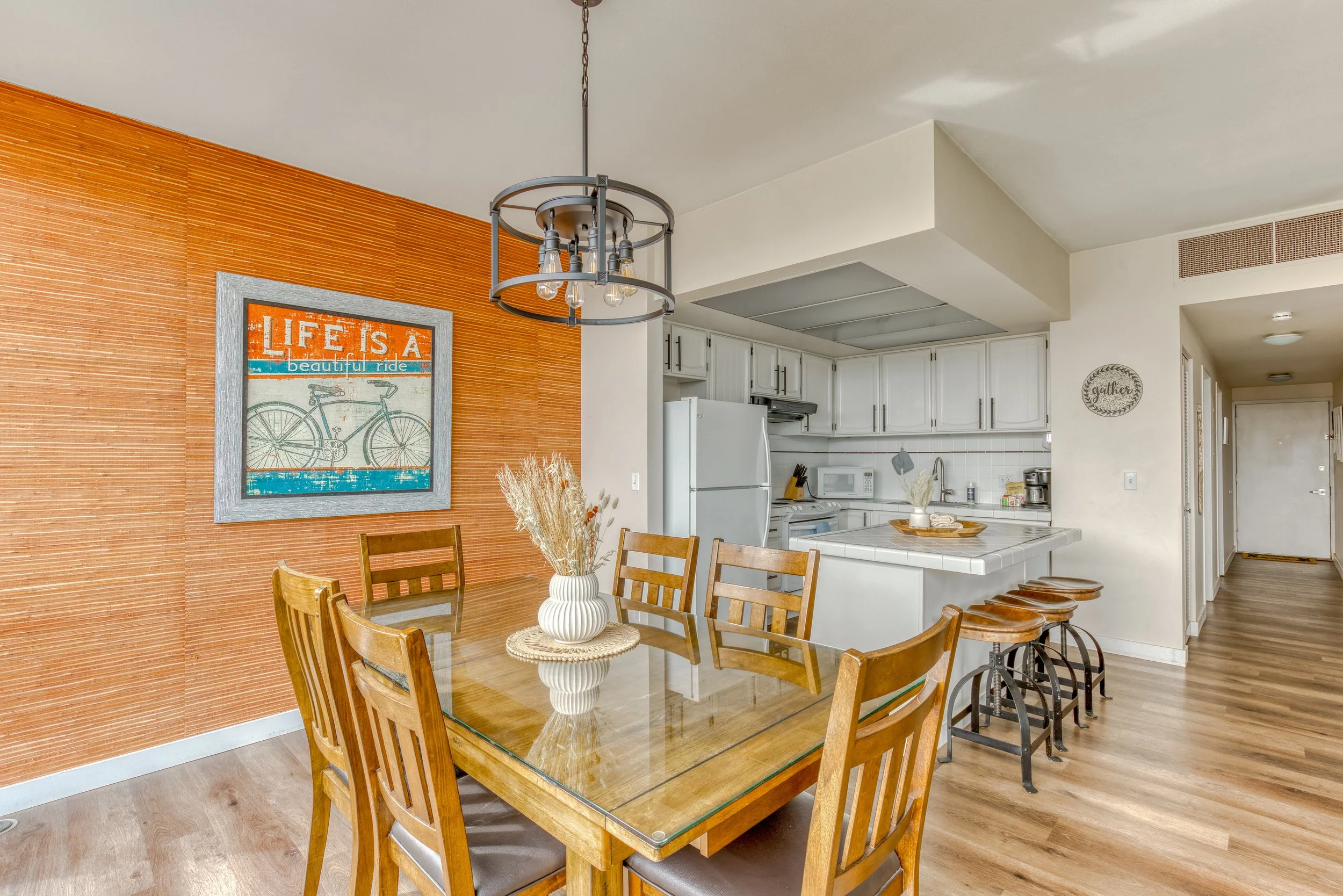 Dining area with wooden table and chairs, decorative orange wall with bicycle artwork, hanging light fixture, and adjacent kitchen with white cabinets and appliances.