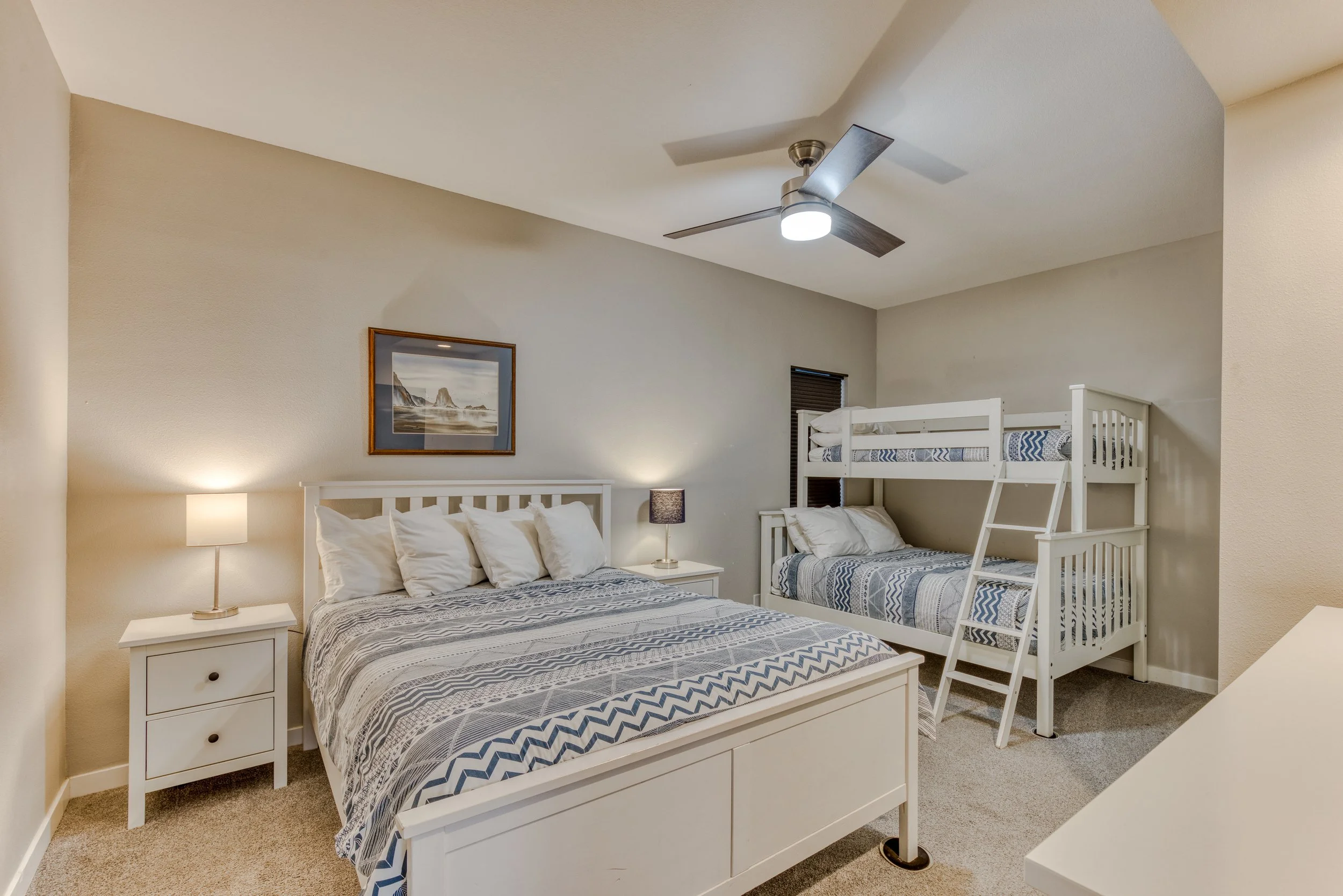 Bedroom with a queen bed and bunk bed, both with blue and white bedding, two side tables with lamps, a ceiling fan, and a landscape painting on the wall.