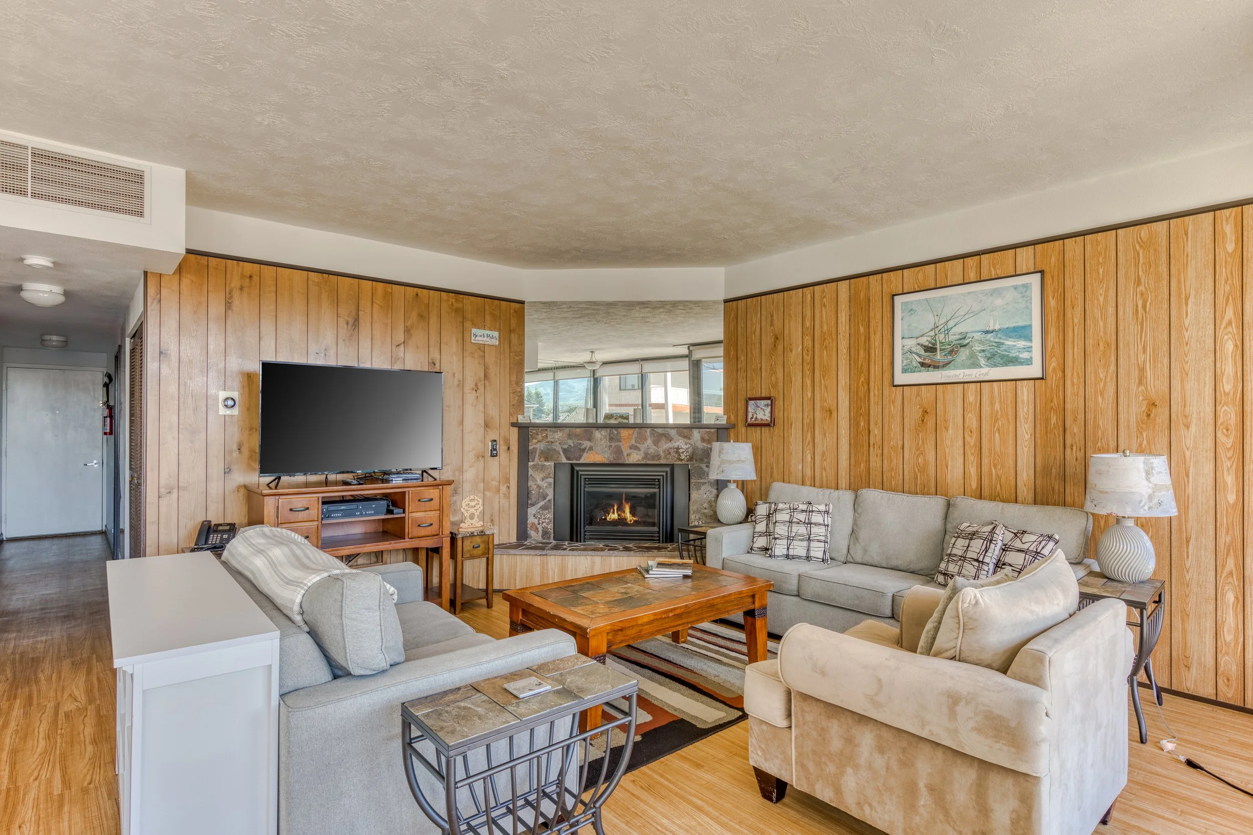 Cozy living room with wood paneling, a fireplace, and a flat-screen TV on a stand. Two sofas and an armchair are arranged around a wooden coffee table. Decor includes a nautical-themed painting and lamps.