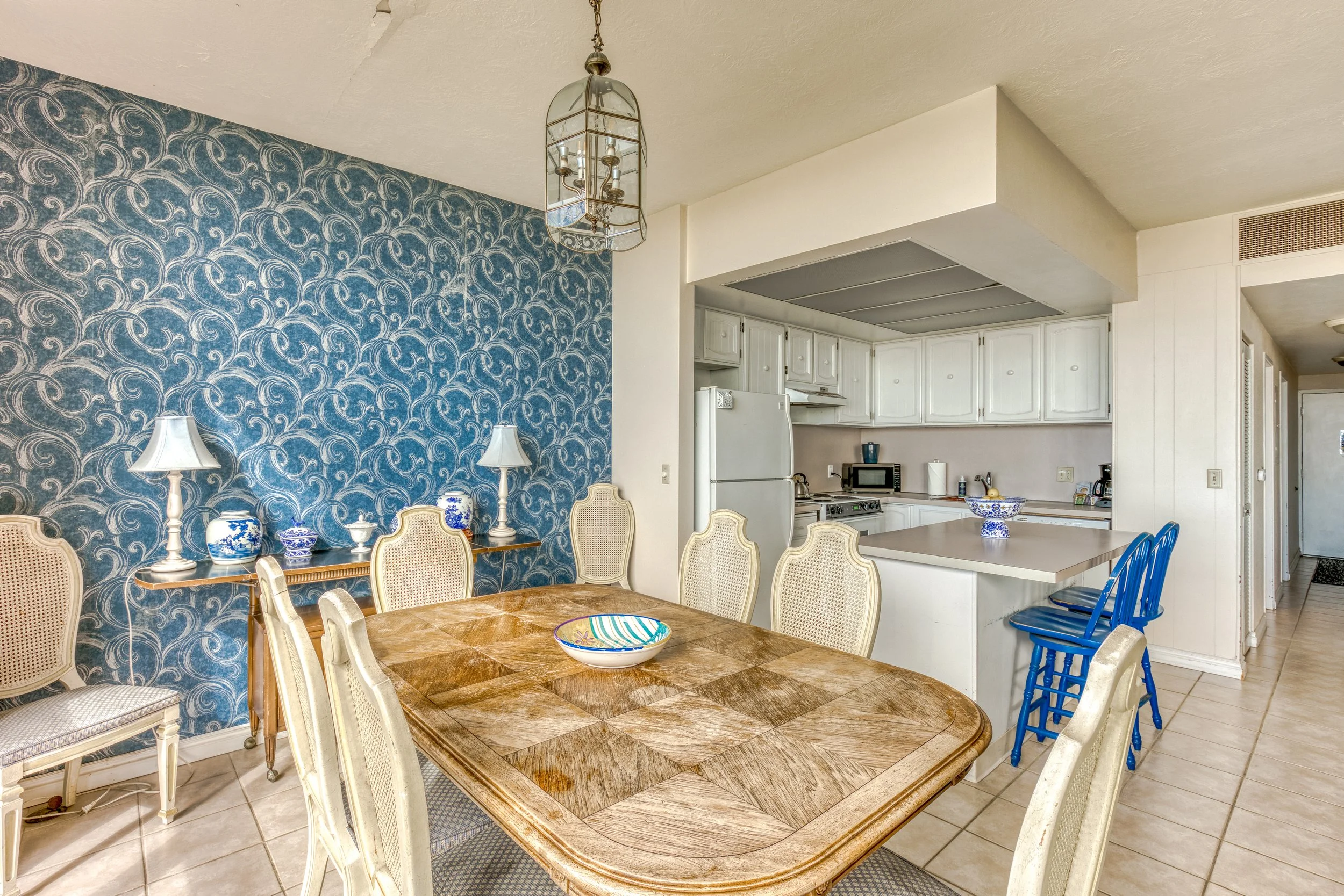 Dining room and kitchen with wooden table, chairs, blue patterned wall, pendant light, and white cabinetry featuring a counter with blue stools.