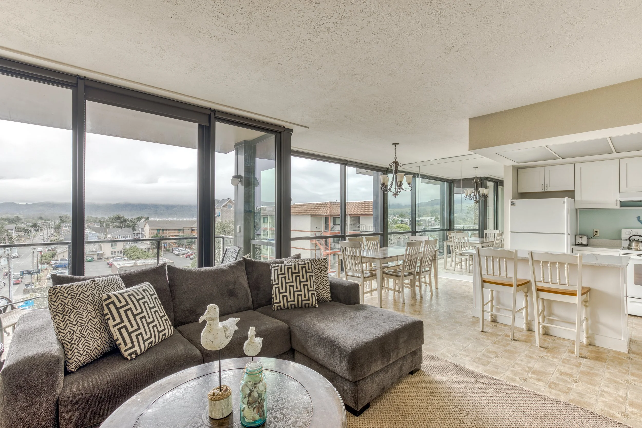 Bright open-concept living and dining area with large windows, gray sectional sofa, decorative pillows, round coffee table with seagull ornaments and jars, white kitchen cabinets, dining table, chairs, and view of buildings and hills outside.