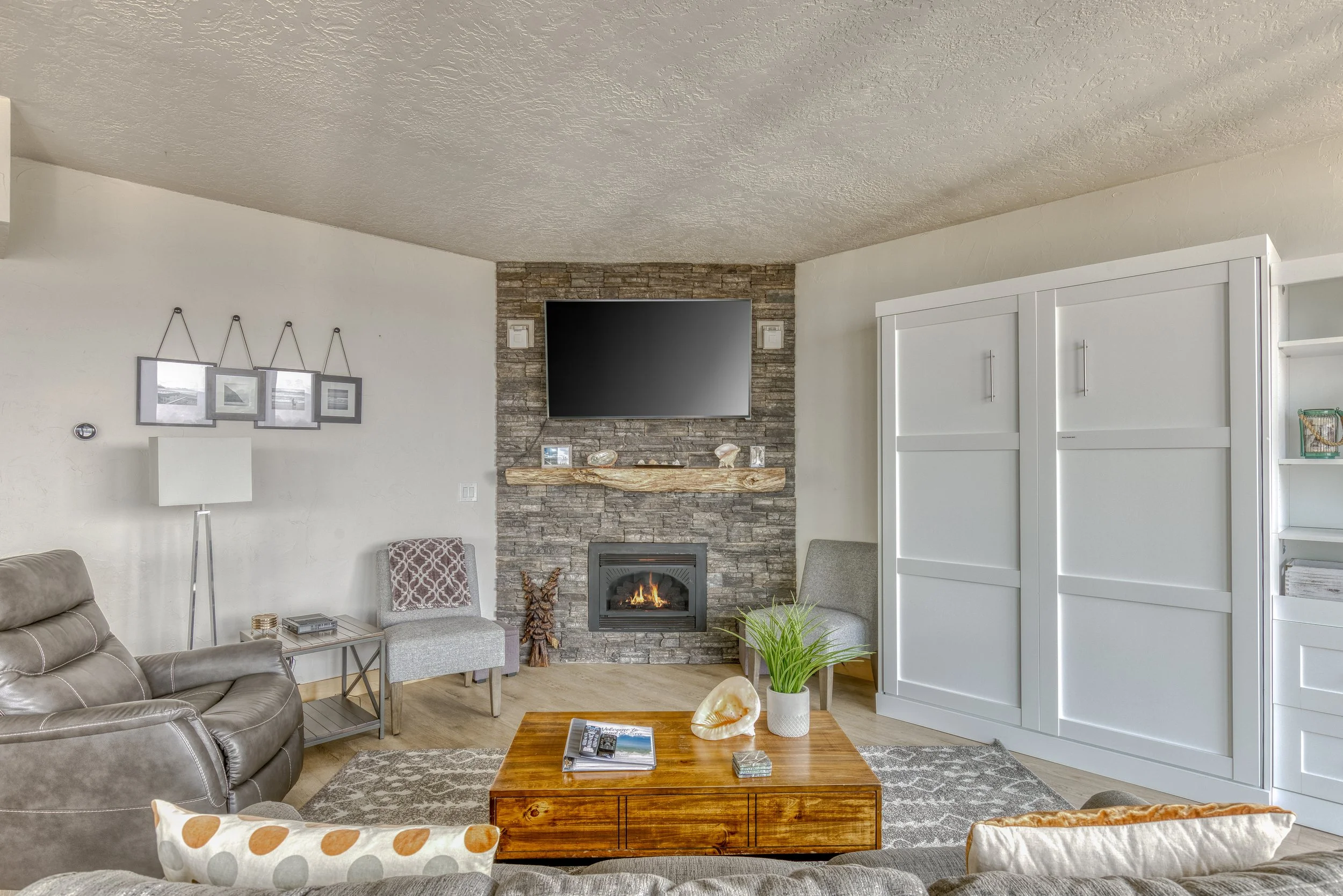 Modern living room with a leather recliner, stone fireplace, wall-mounted TV, wooden coffee table, pillows, and decor accents.