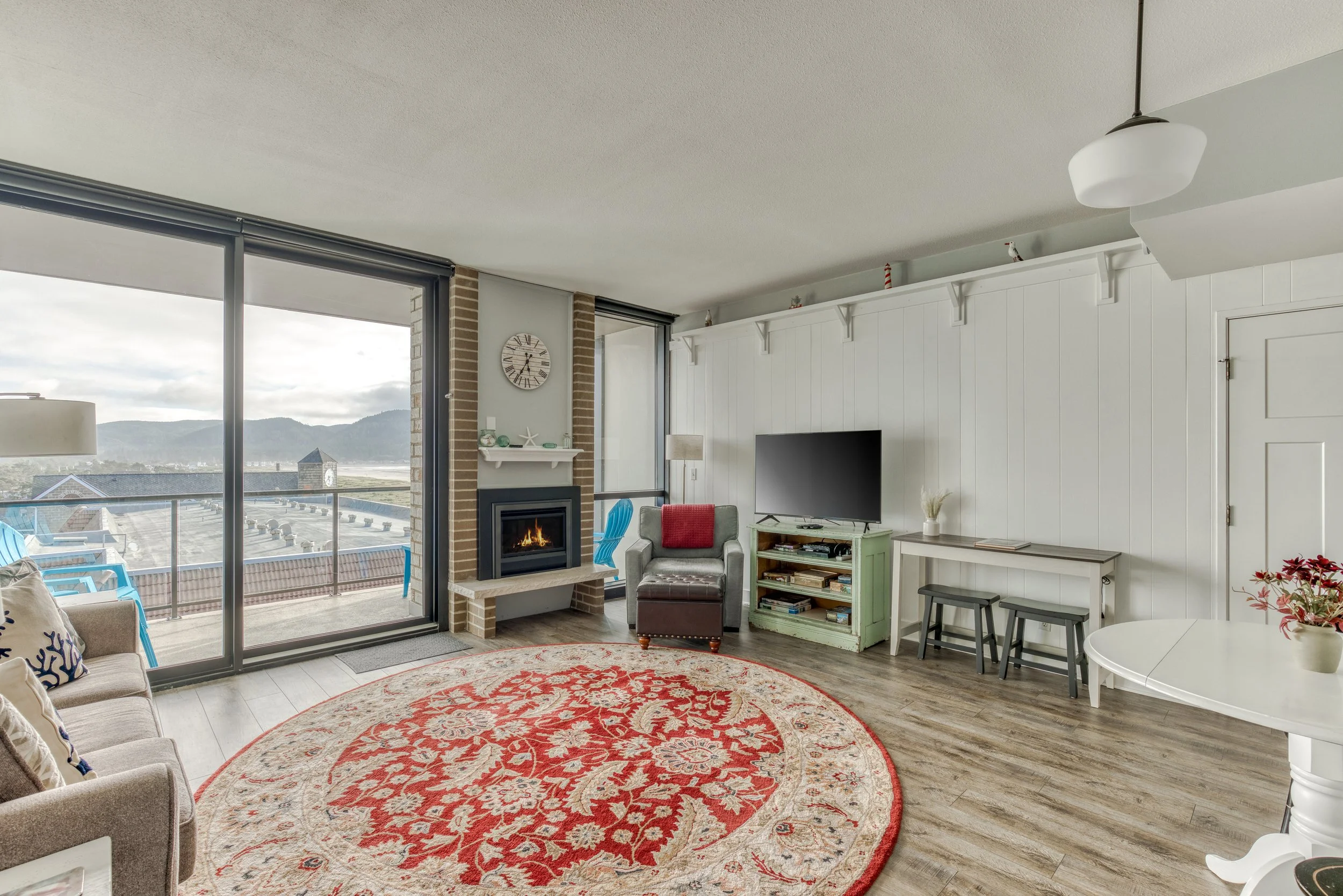 Cozy living room with large windows, a fireplace, and a view of mountains. Features a red patterned rug, sofa, armchair, TV, and a small table with stools. Outdoor balcony visible through glass doors.