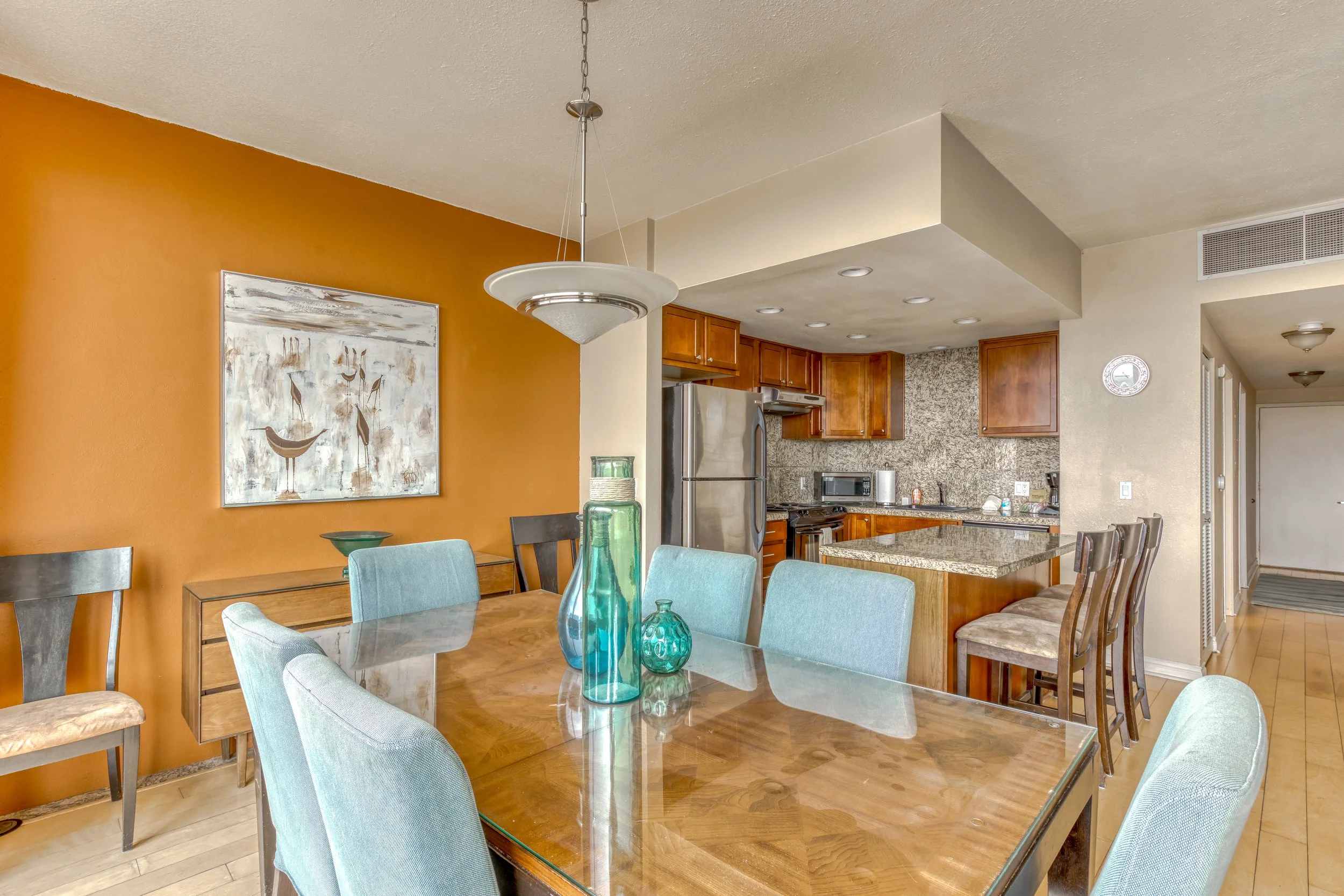 Dining room with glass-top table, aqua chairs, and kitchen view in modern home.