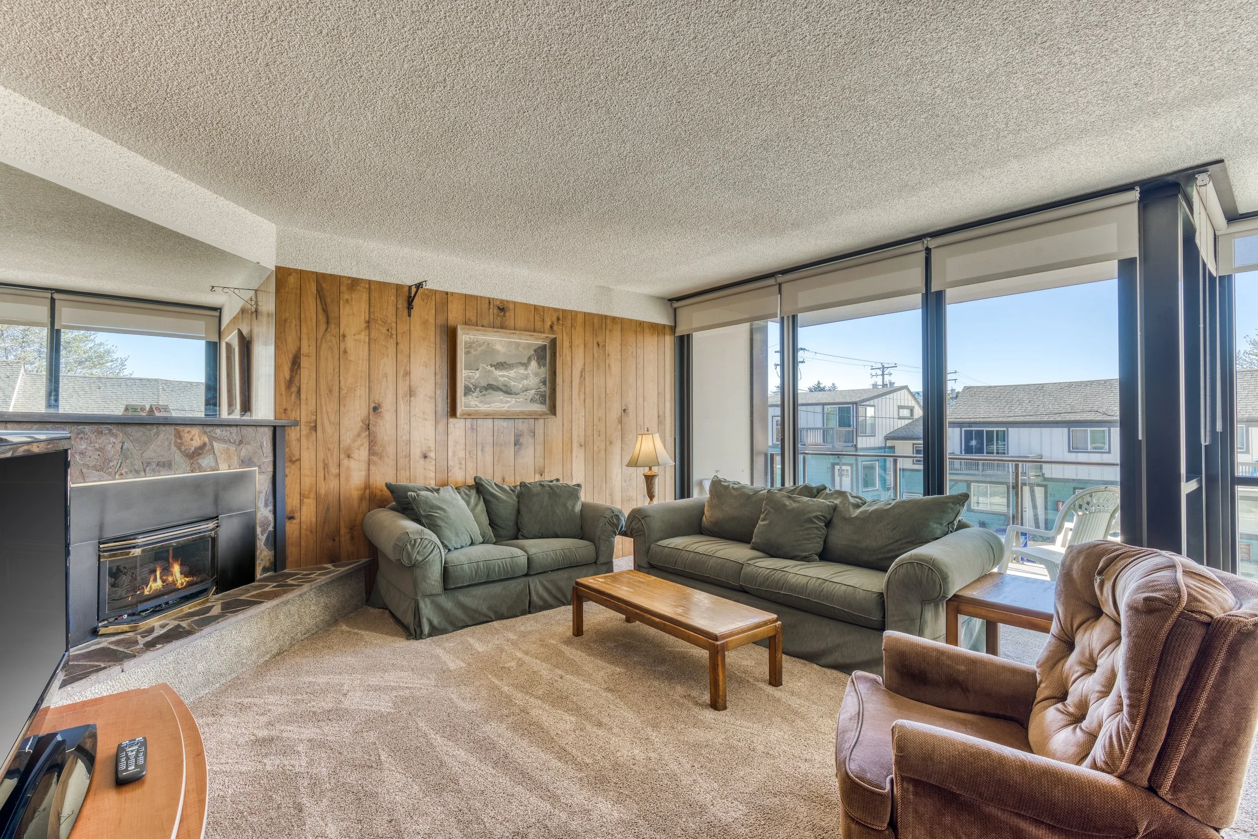 Cozy living room with two green sofas, a brown armchair, wooden coffee table, brick fireplace, and large windows with outdoor view.