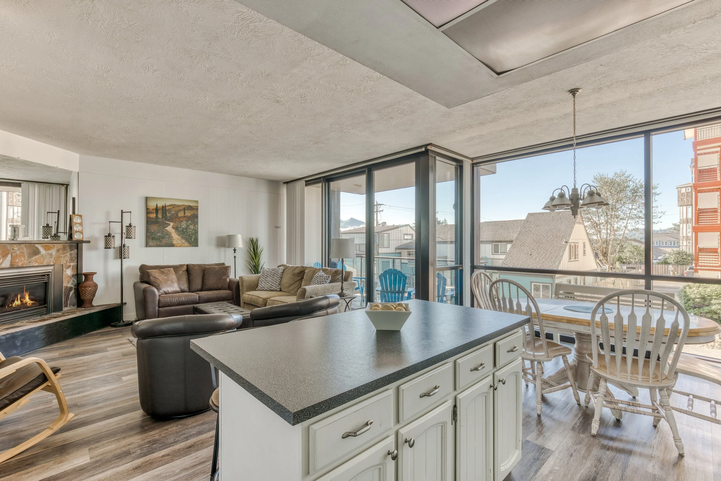 Open-concept living room and dining area with modern decor. Features brown leather and beige fabric sofas, a rocking chair, a fireplace, large windows with a view outside, an island with a countertop, and a wooden dining table with chairs. Hardwood flooring is present.