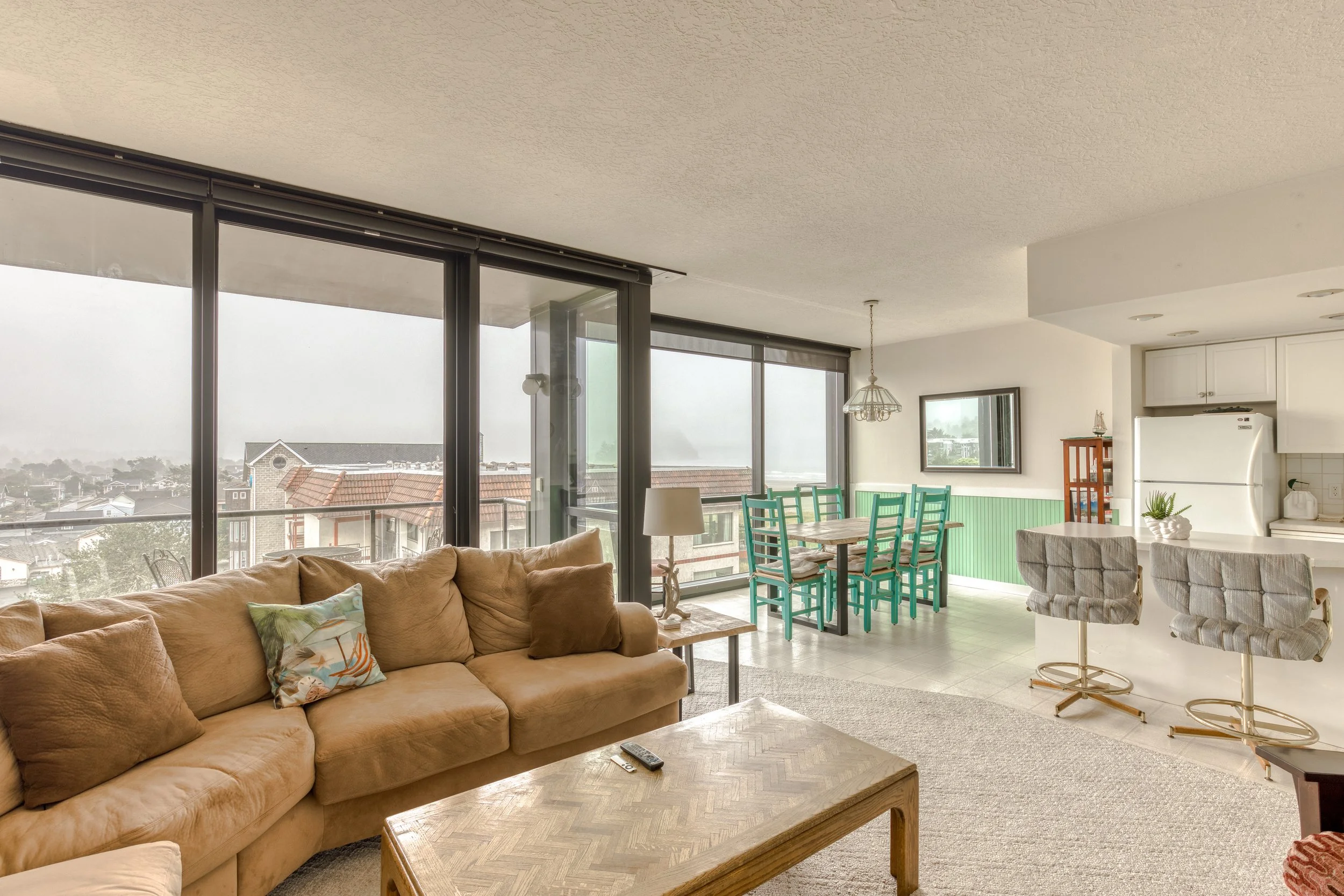 A bright living room with large windows, featuring a beige sectional sofa, a wooden coffee table, a dining area with green chairs, and a kitchen with white cabinets. The space overlooks a balcony with a city view.