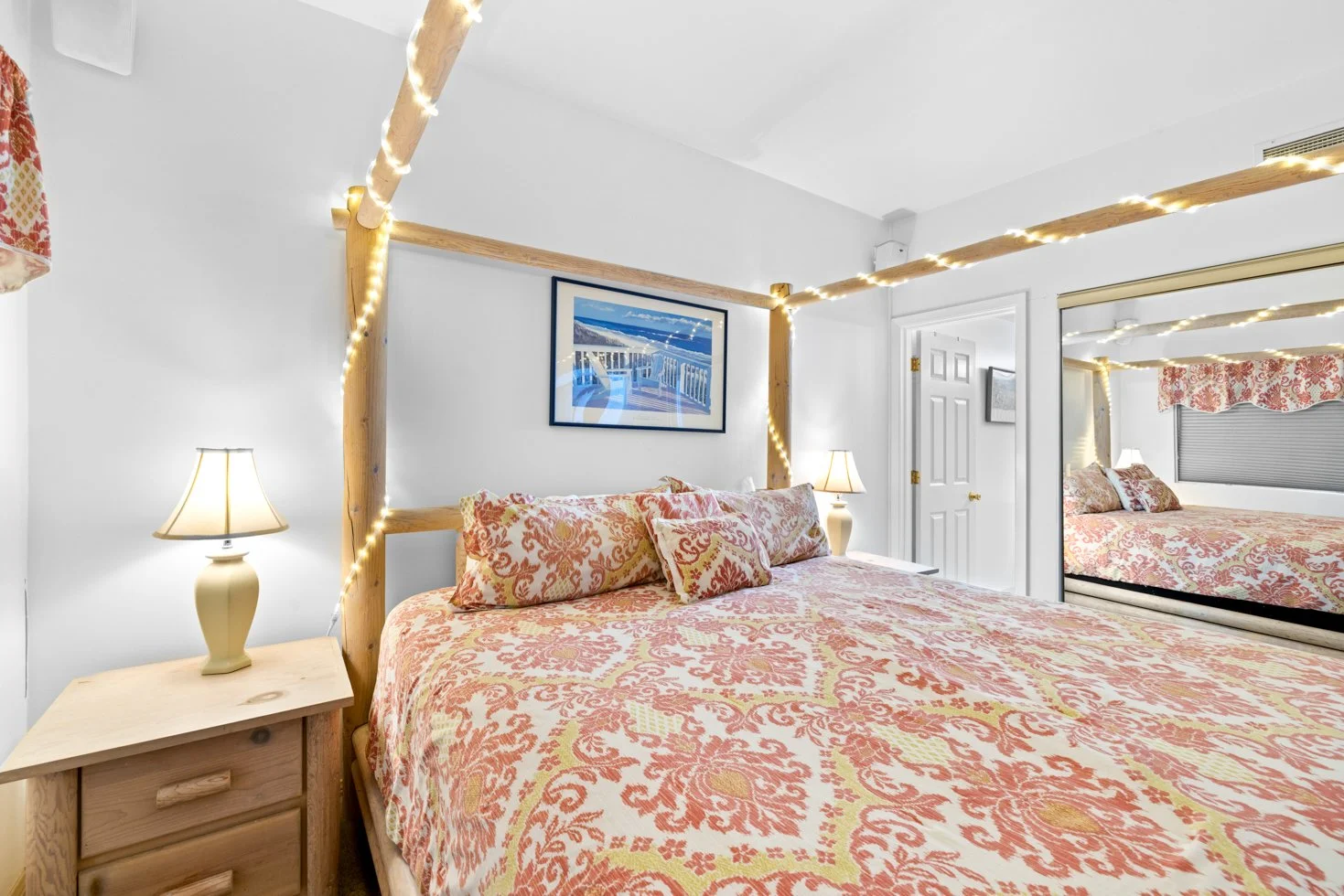 Cozy bedroom with a large bed adorned with a patterned duvet, fairy lights on a wooden canopy frame, two bedside lamps, a wooden nightstand, and a mirror reflecting the room. A framed picture hangs above the bed.