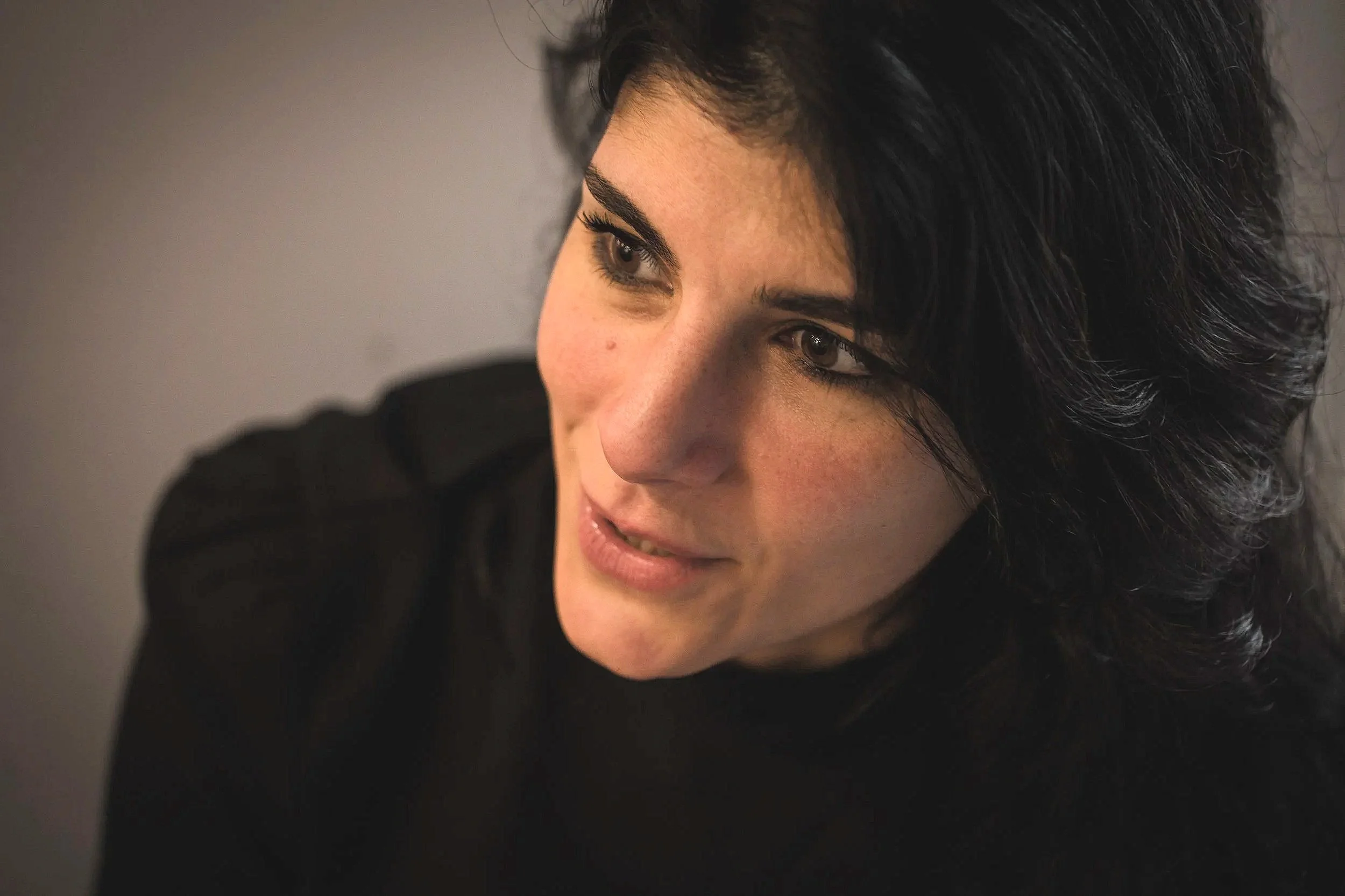 Nahaufnahme eines Frauenkopfes mit dunklen, gewellten Haaren, die leicht nach vorne fallen, und einem schwarzen Oberteil. Sie blickt leicht nach oben und lächelt sanft.