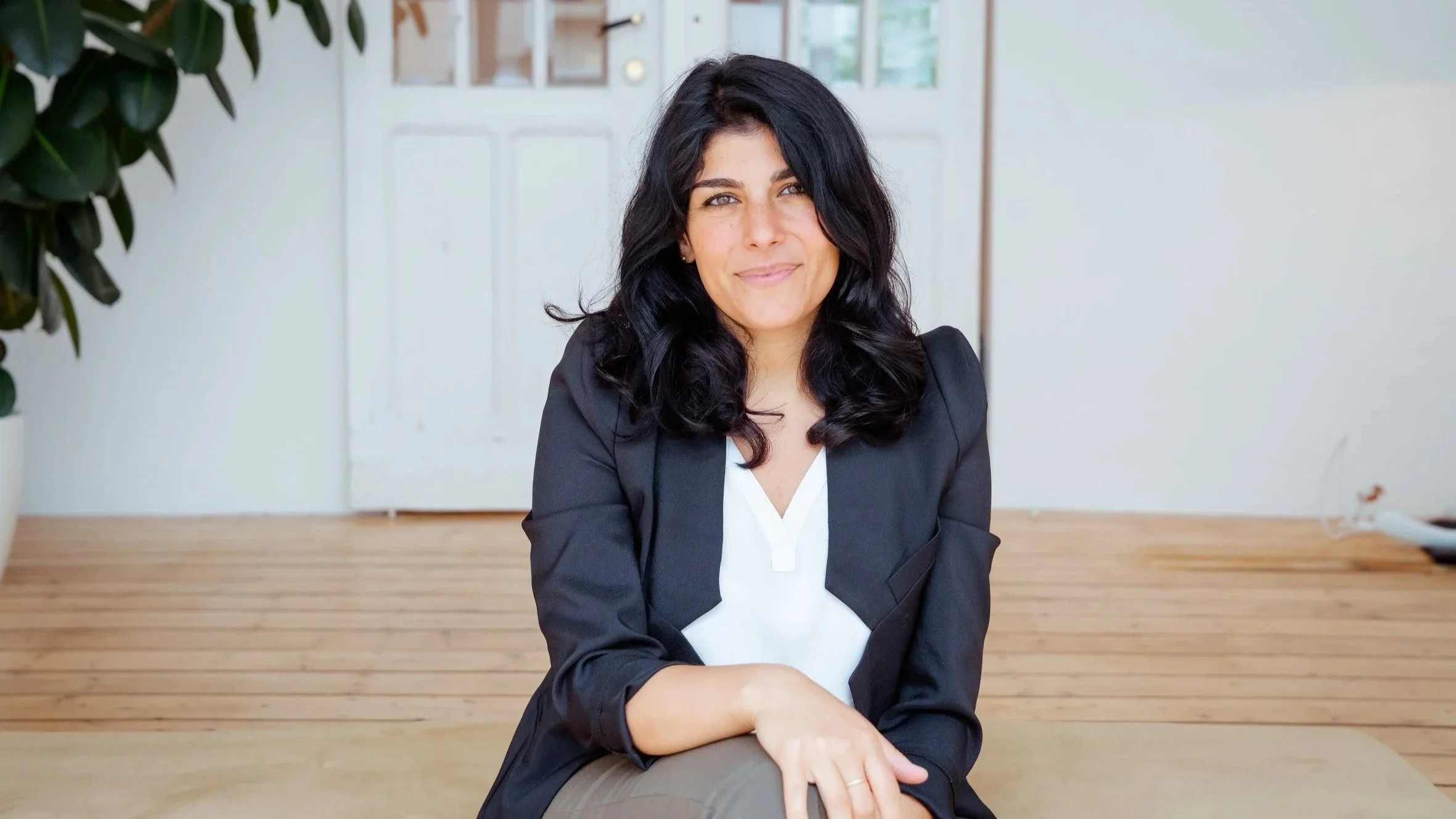 Eine Frau mit dunklen, lockigen Haaren, trägt eine schwarze Blazer und sitzt an einem Holztisch in einem hell erleuchteten Raum mit Holzfußboden und weißem Tür im Hintergrund.