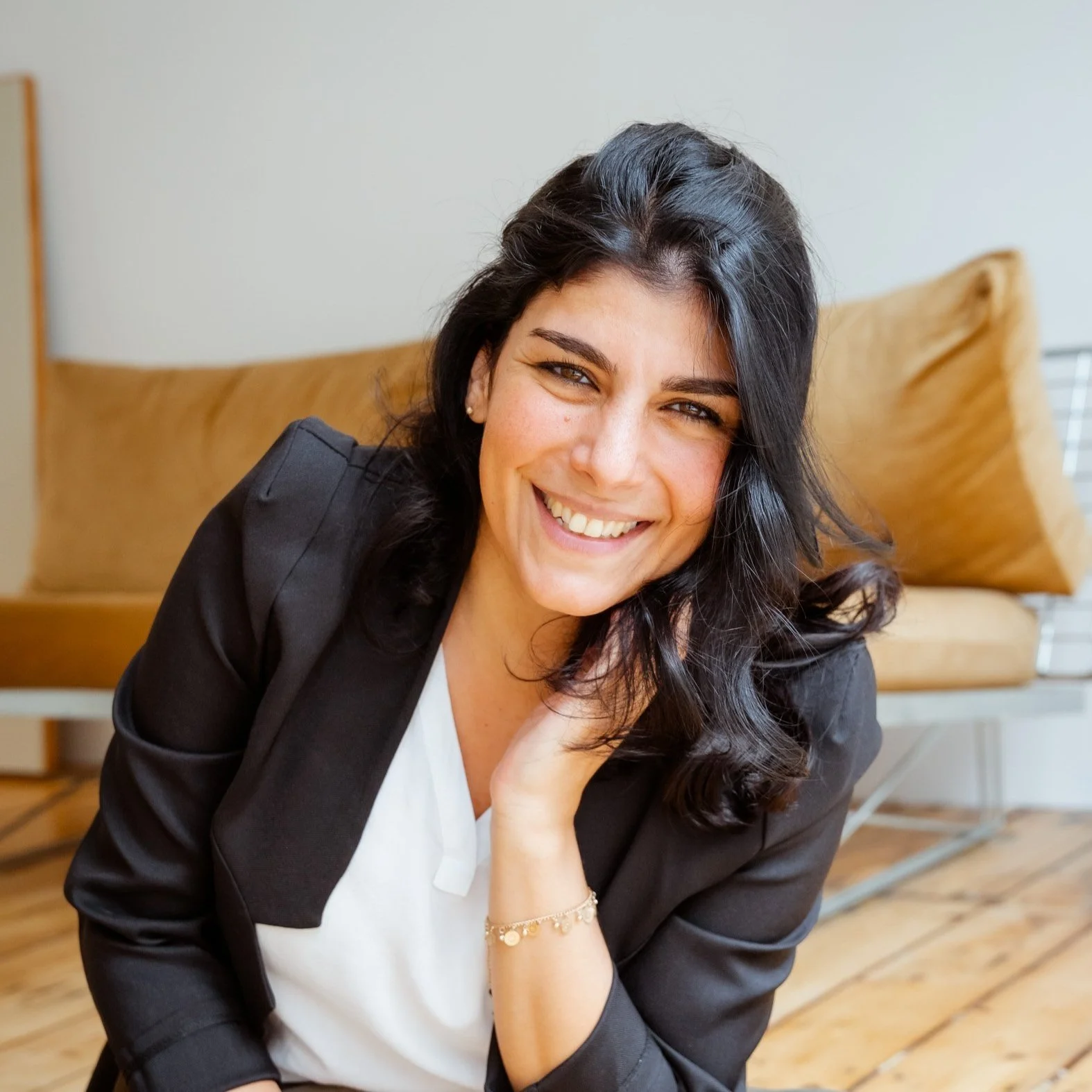 Lächelnde junge Frau mit dunklen Haaren in einem schwarzen Blazer und weißem Hemd auf einem Holzboden, im Hintergrund braune Couch und graue Wand.