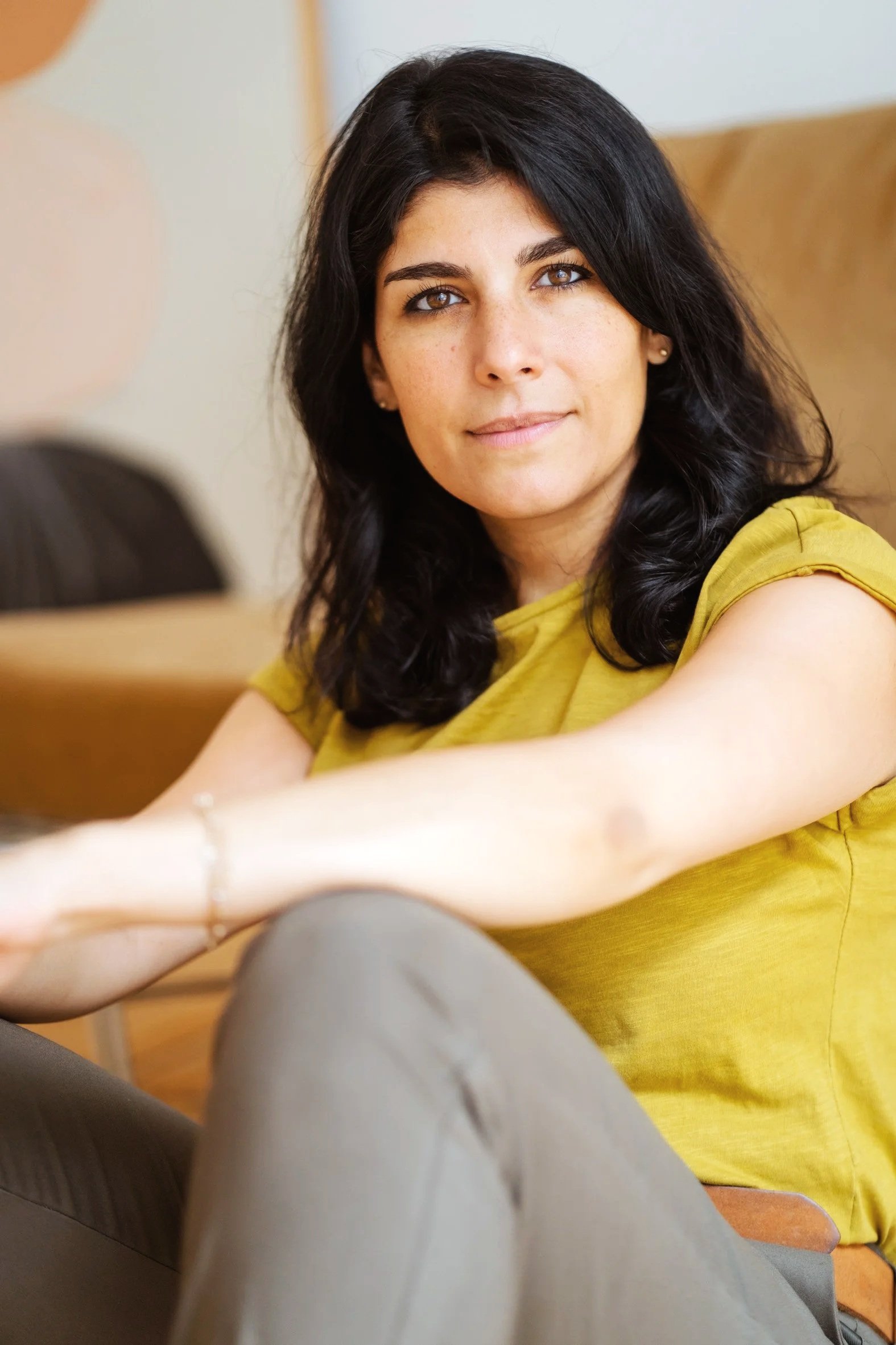 Frau mit dunklen Haaren und gebräunter Haut trägt ein gelbes Oberteil, sitzt auf dem Boden angelehnt an einer Couch und schaut direkt in die Kamera, im Hintergrund eine weiße Wand und eine braune Couch.