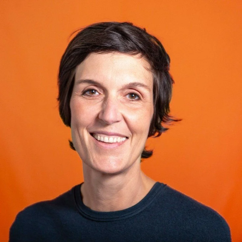 Ein lachender Frau mit kurzen dunklen Haaren vor orangefarbenem Hintergrund.