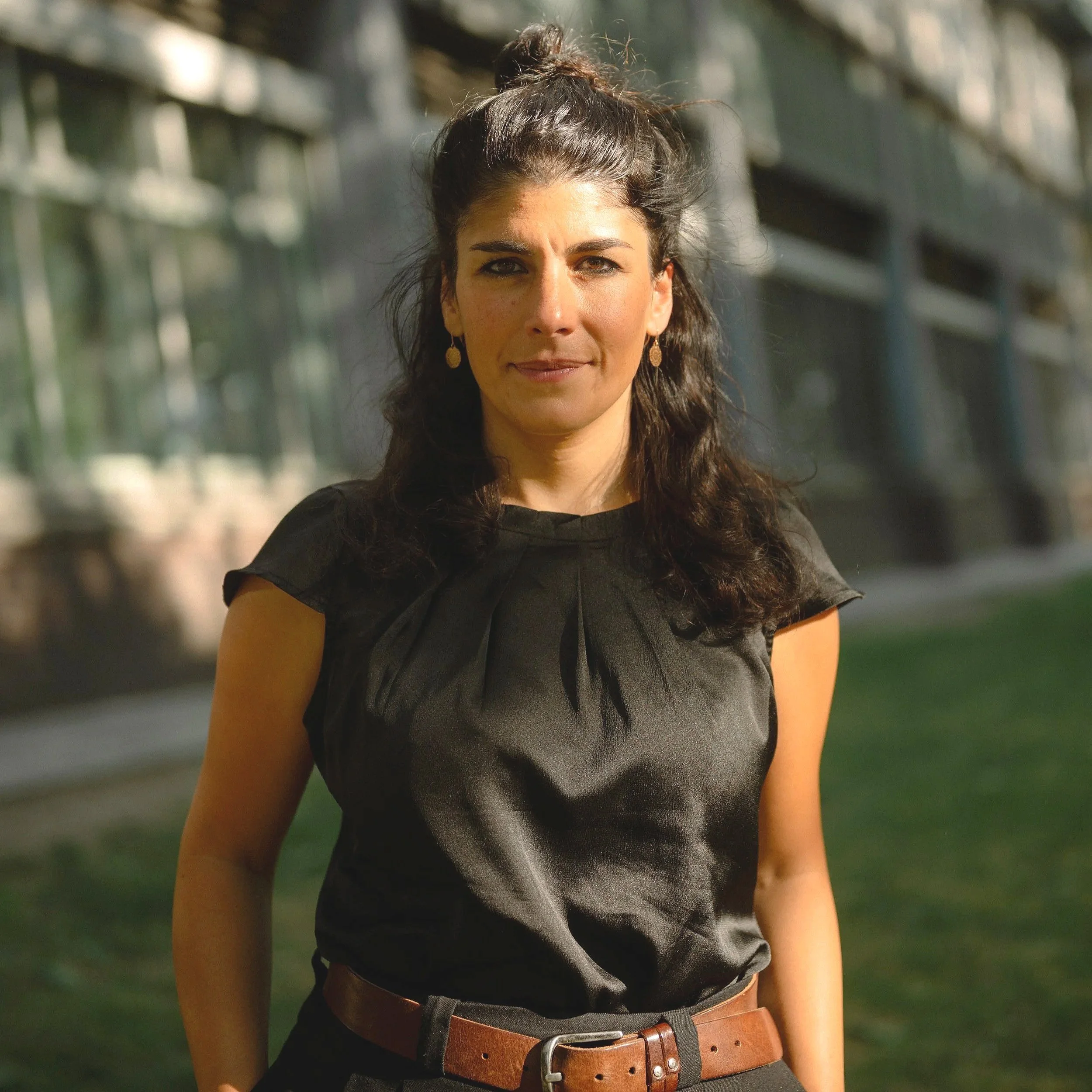 Eine Frau mit dunklen, gewellten Haaren in einem schwarzen Top steht im Freien, im Hintergrund ein modernes Gebäude und grüne Rasenfläche.