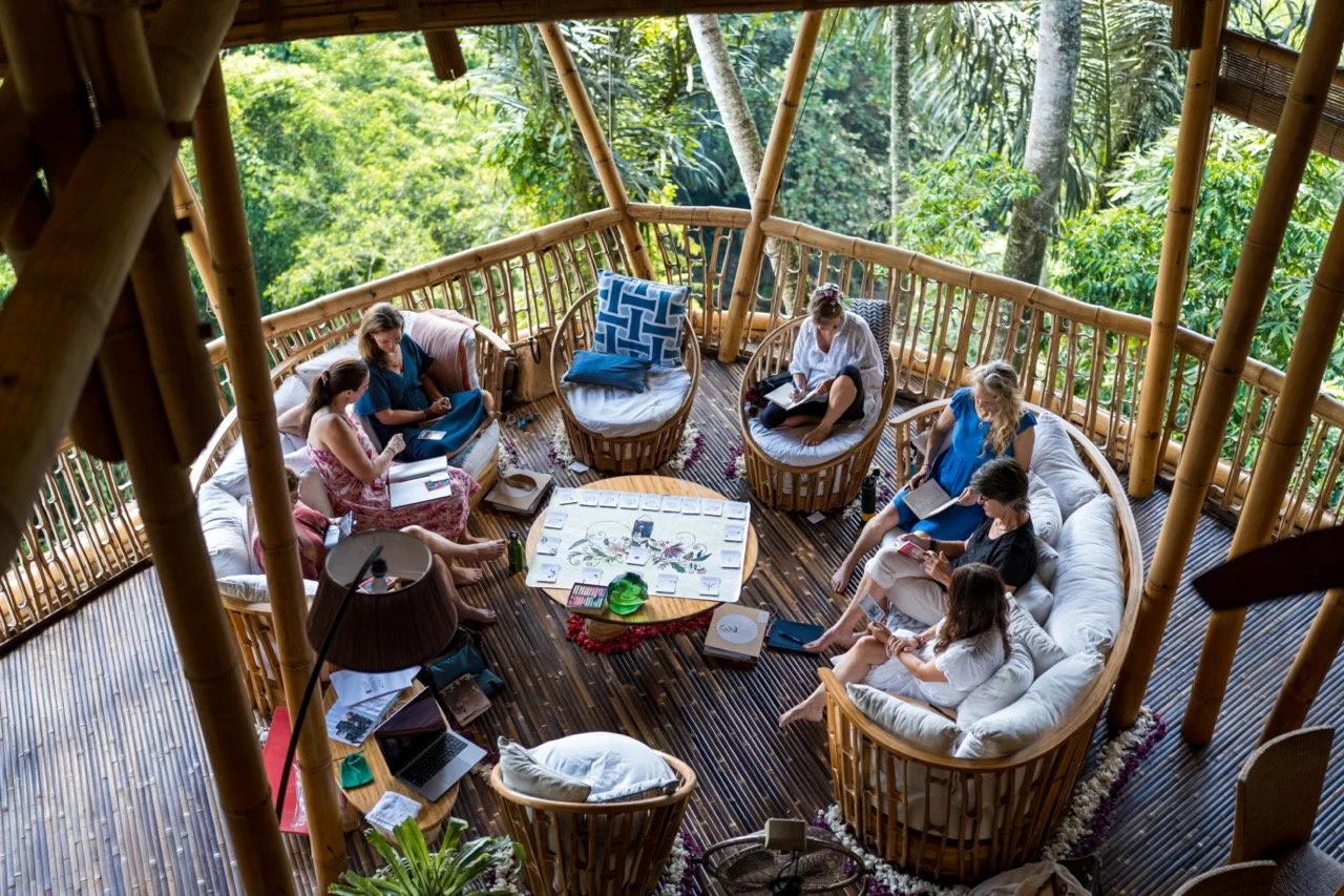 A group of people sitting in a circular arrangement on bamboo furniture inside a treehouse, surrounded by lush green trees, engaging in a meeting or discussion with notebooks, tablets, and a board game on a central table.