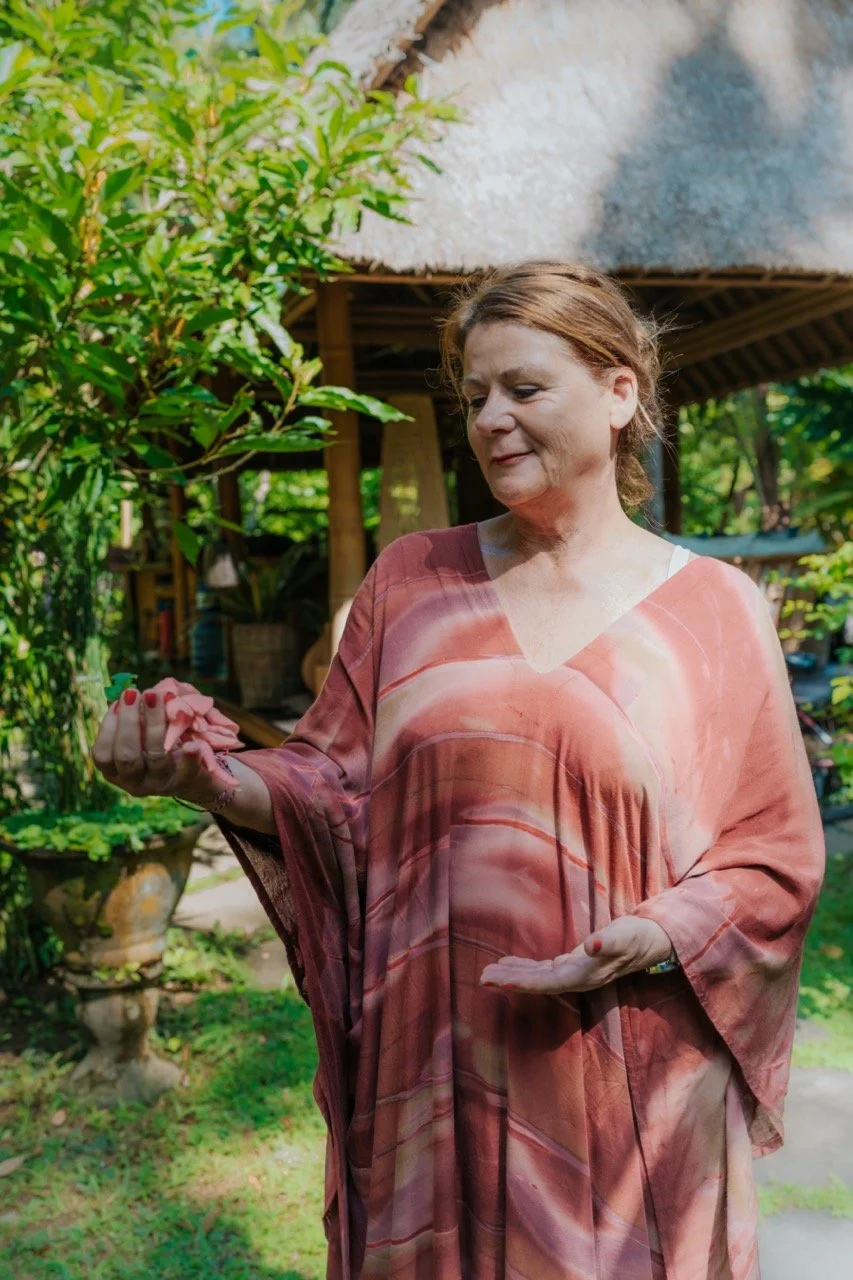 A middle-aged woman with reddish-brown hair wearing a flowing, patterned dress looking at an object in her hand outdoors, surrounded by lush greenery and a thatched roof structure in the background.