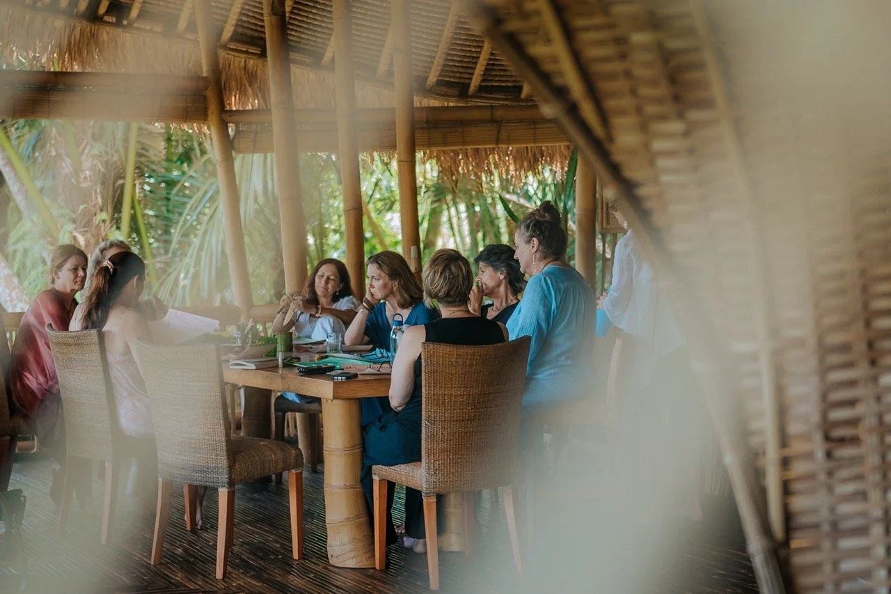 Group of women sitting around a wooden table engaged in discussion inside a bamboo and thatched-roof structure with lush greenery in the background.