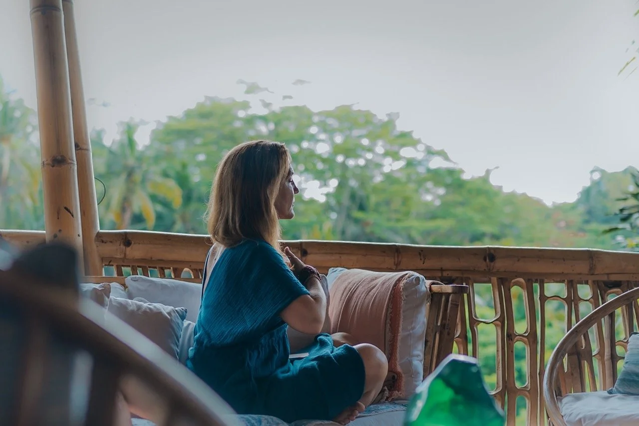 Woman sitting cross-legged on a bamboo porch, gazing at green foliage outside, with cushions and a glass bottle nearby.