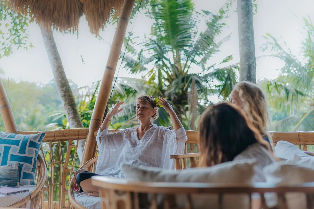 Three women relaxing on a bamboo porch surrounded by tropical trees, one meditating with eyes closed and hands raised, others sitting and listening.
