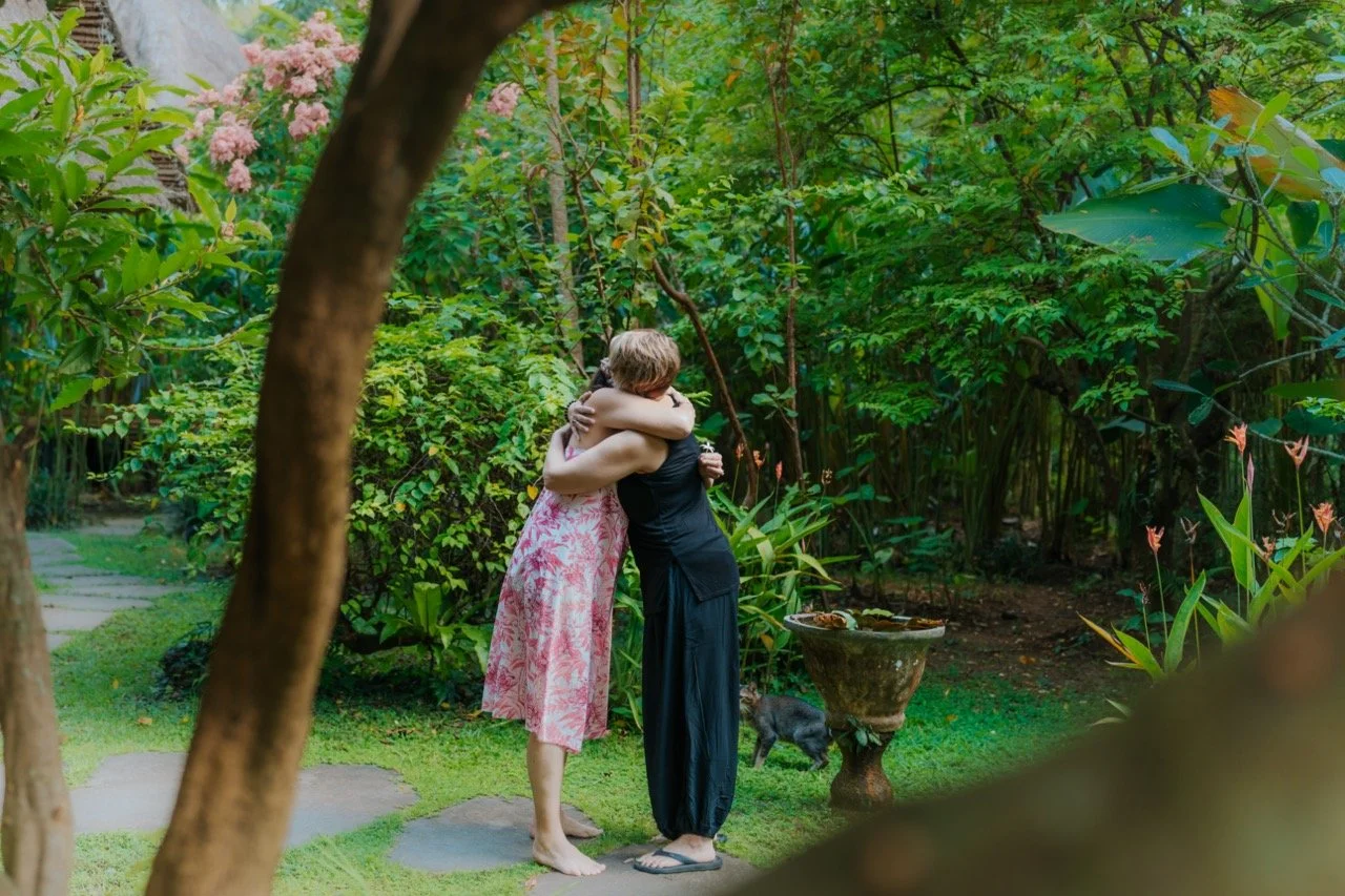 Two women hugging in a lush garden surrounded by green plants and trees, with a small black piglet on the ground nearby.