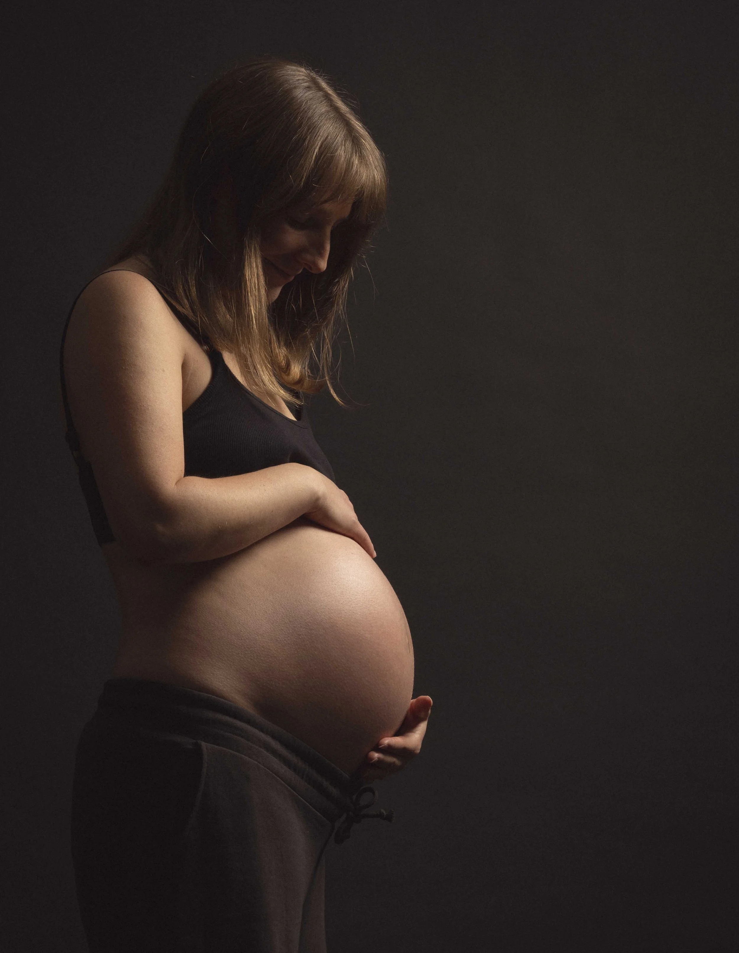 A pregnant woman with long hair, wearing a black sleeveless top and dark sweatpants, looking down at her belly and smiling against a dark background.