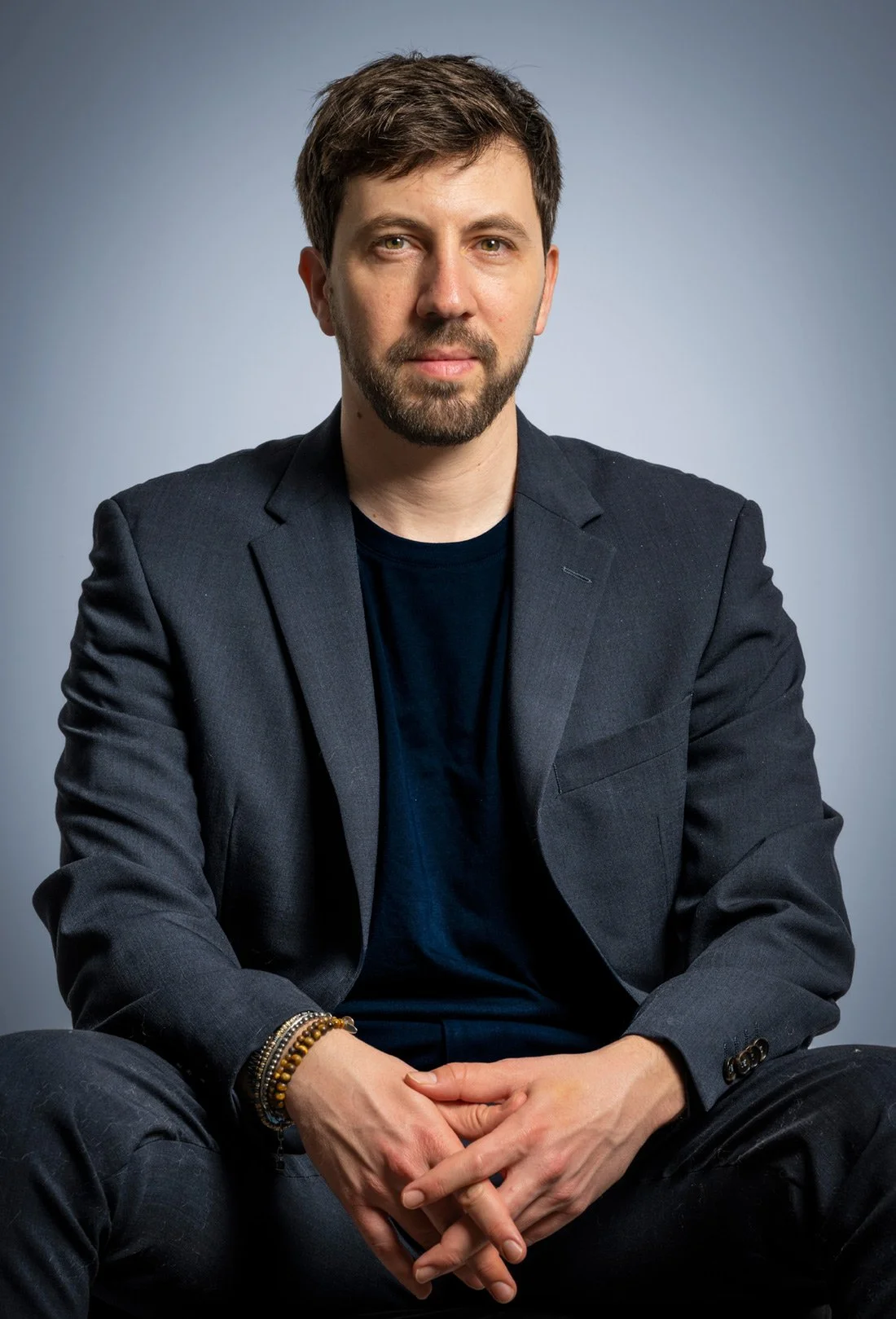 A man with brown hair and beard wearing a black blazer and black shirt, sitting against a gray background.