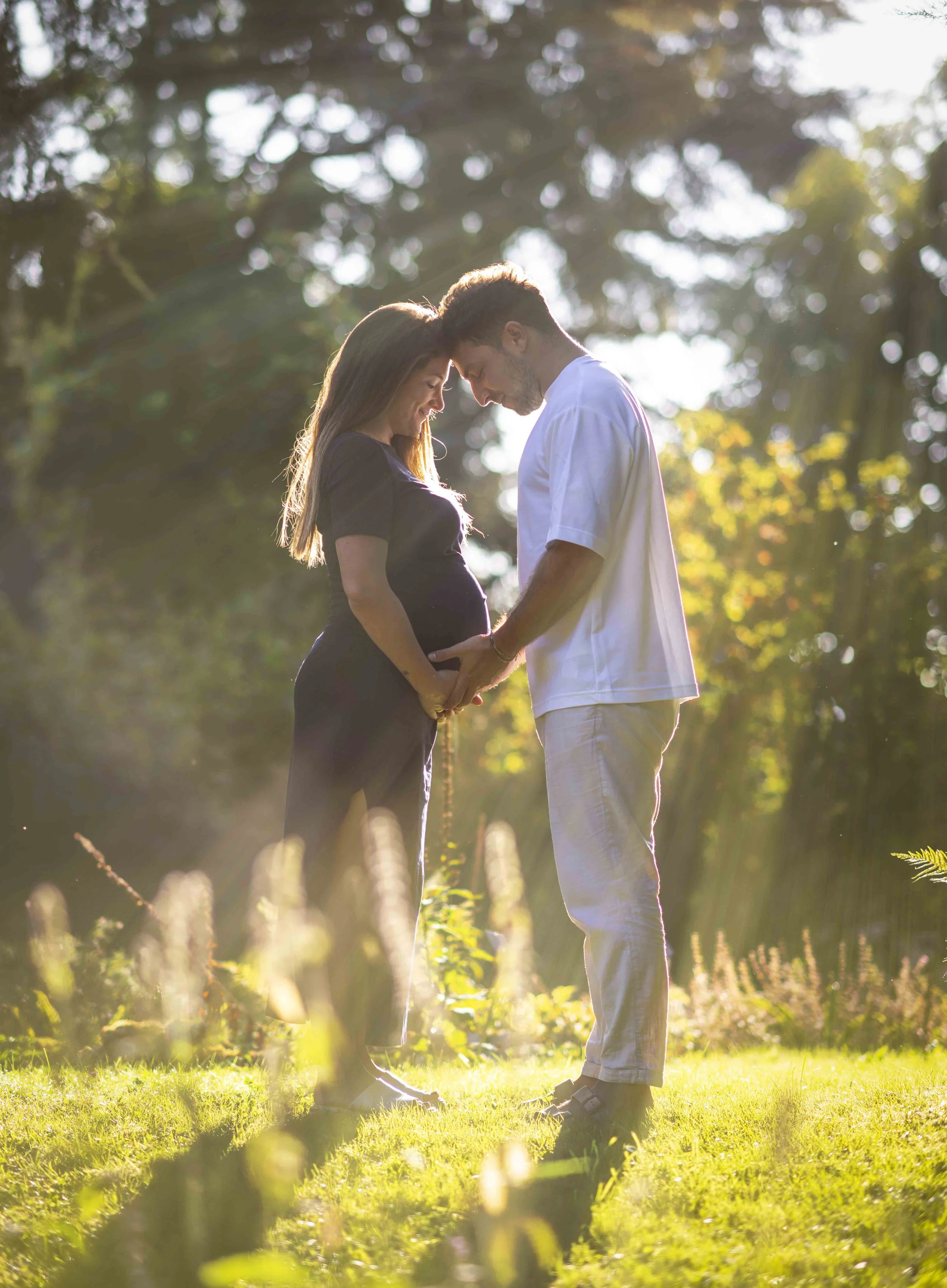 A pregnant woman and a man stand closely outdoors in a sunlit garden, gently touching her baby bump with their foreheads touching.