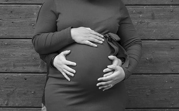Pregnant woman cradling her belly with both hands in front of a wooden wall.