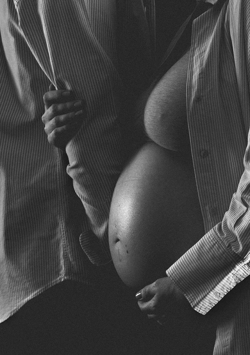 Close-up of a pregnant woman in a partially unbuttoned striped shirt, showing her bare belly and chest, with her hand resting on her belly.