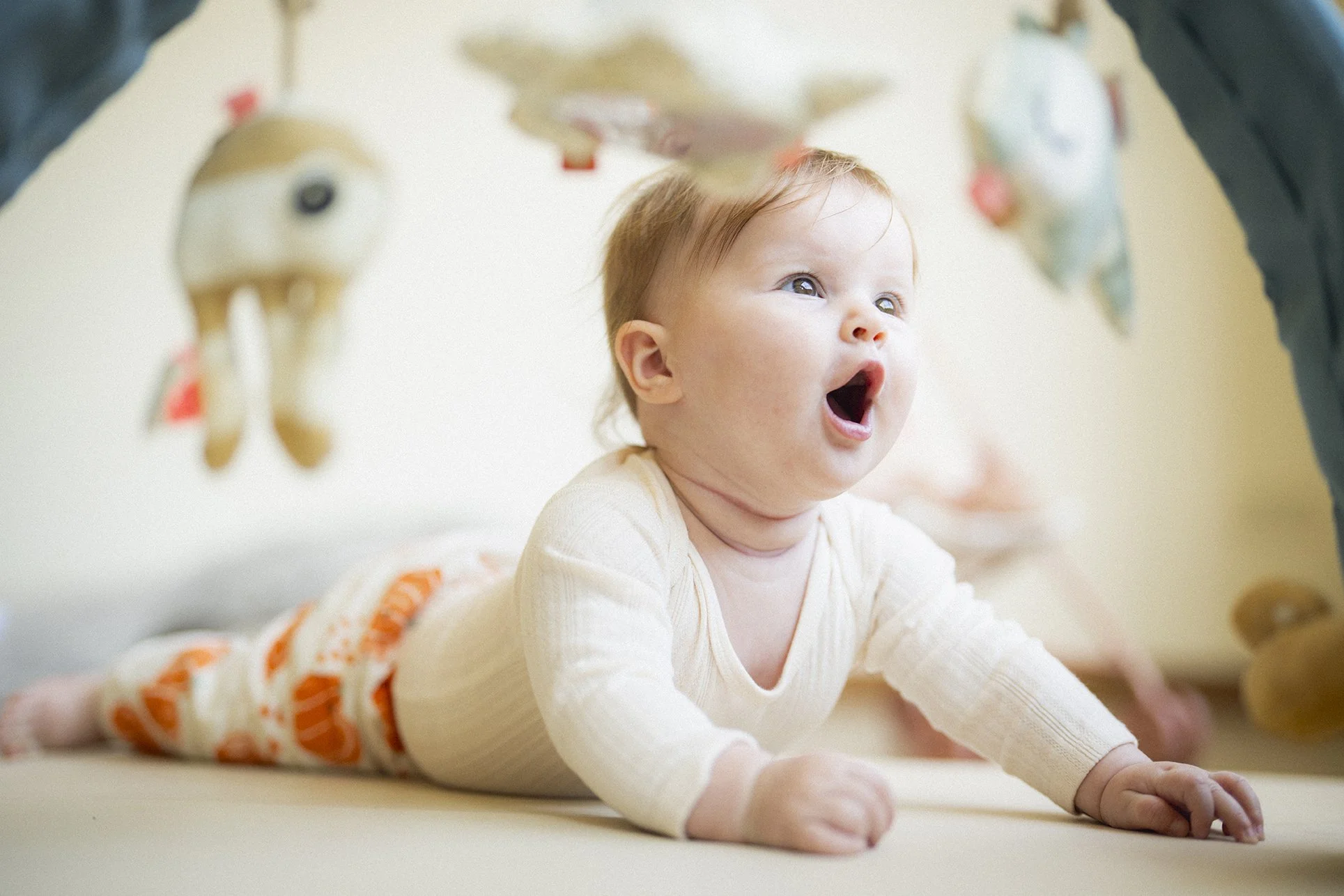 Baby lying on stomach as he looks at a hanging toy mobile above him, with a surprised or excited expression.
