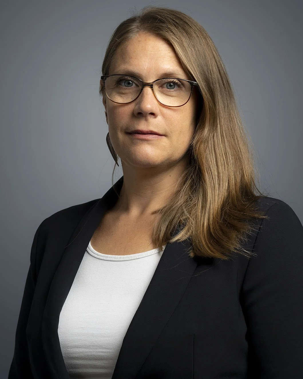 A woman with shoulder-length light brown hair, wearing glasses, a black blazer, and a white top, posing against a gray background.