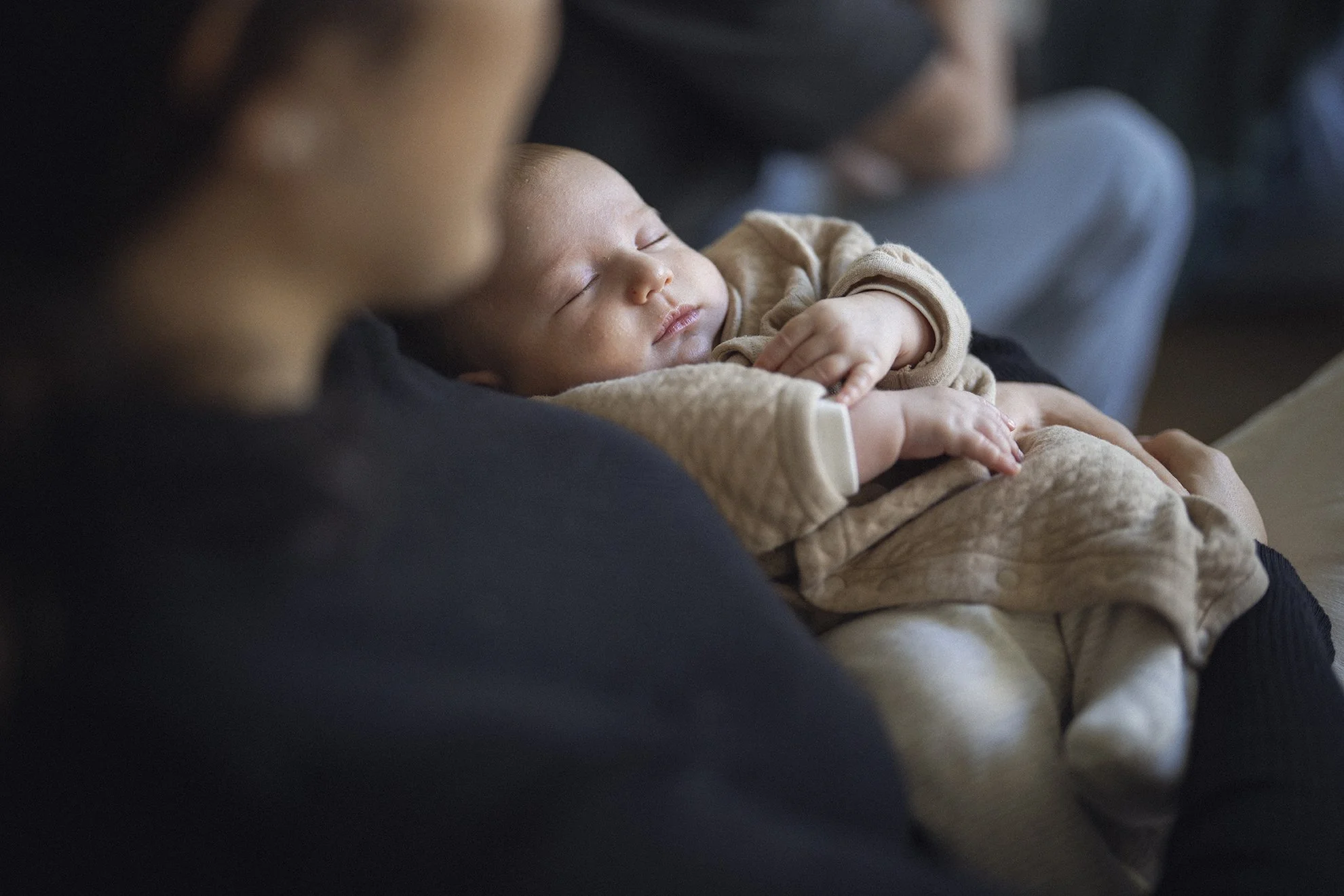 A sleeping baby is peacefully cradled in an adult's arms, with the adult's face slightly blurred in the foreground. The background shows other people sitting and talking.