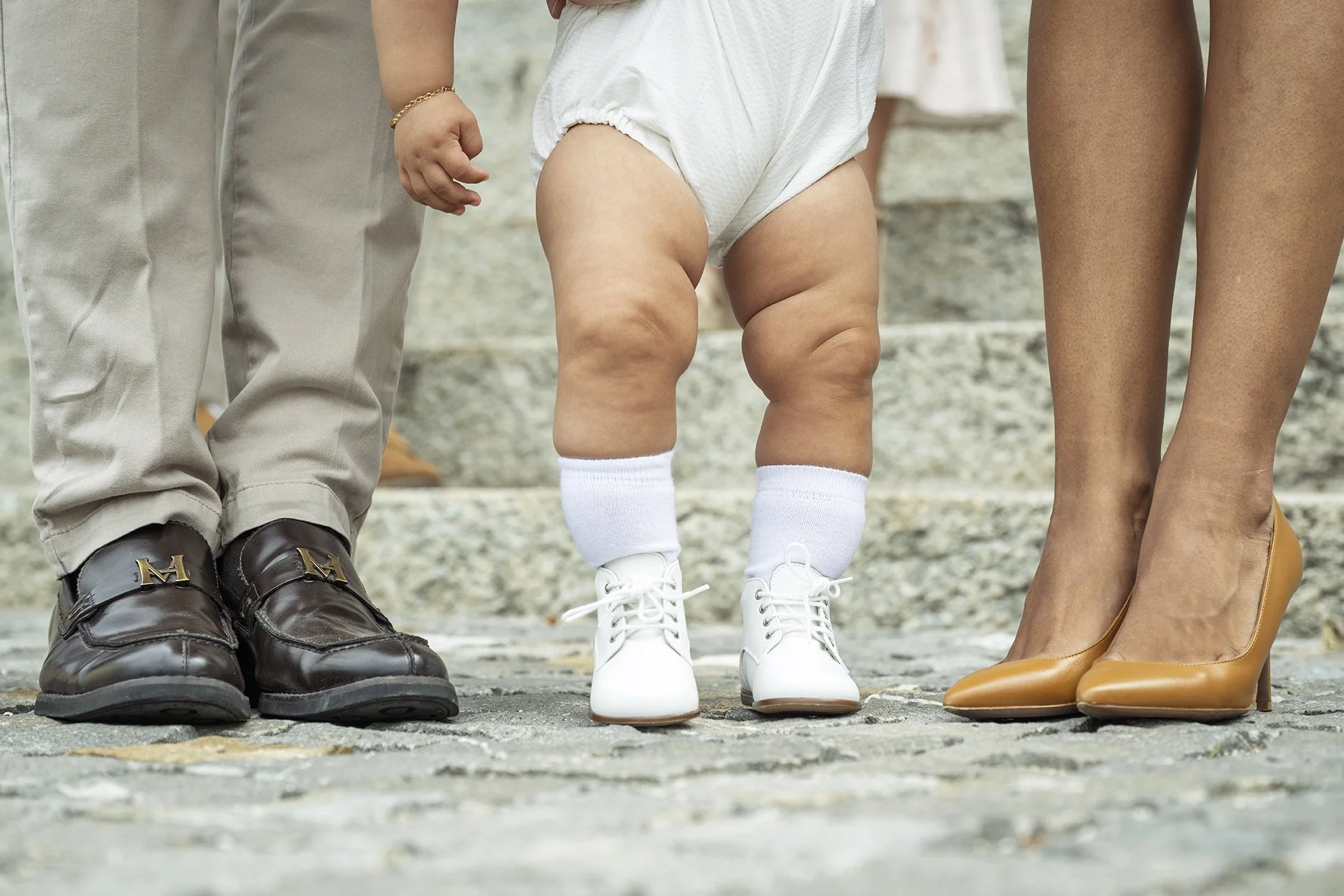 Close-up of three pairs of legs and shoes, with a baby in the middle standing on stone steps. The baby is wearing white shoes, white socks, an oversized white diaper, and has chunky legs. The person on the left is wearing black dress shoes and beige 