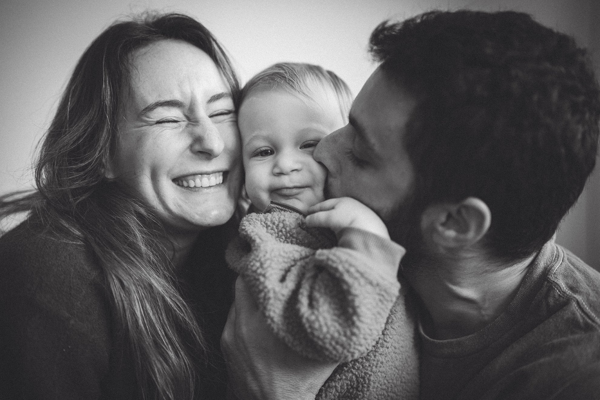 Black and white photo of a smiling woman with long hair, a young child with short hair, and a man with short hair; the man is kissing the child's cheek while the woman holds the child