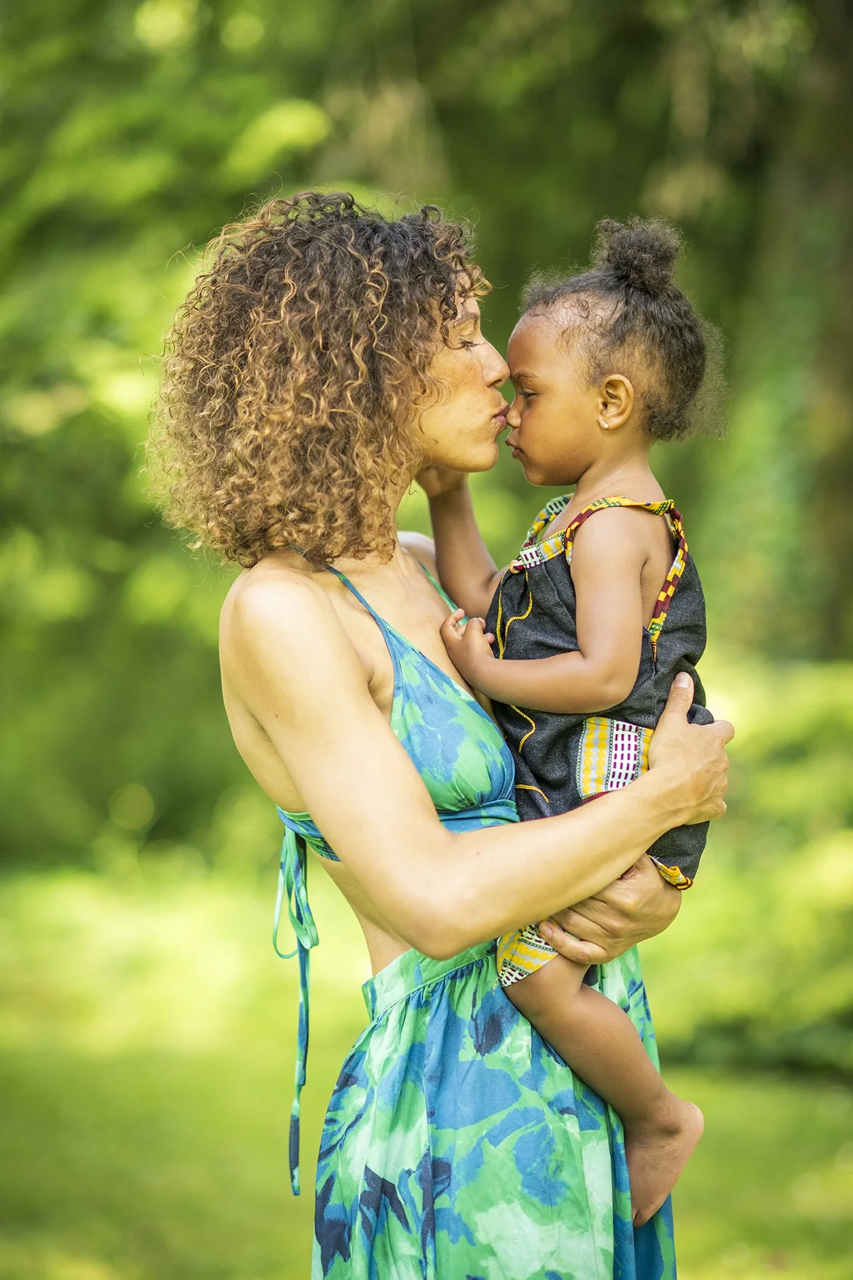 A woman in a blue and green dress holding a young girl with an afro hairstyle in a black dress with colorful accents, outdoors with green foliage in the background.
