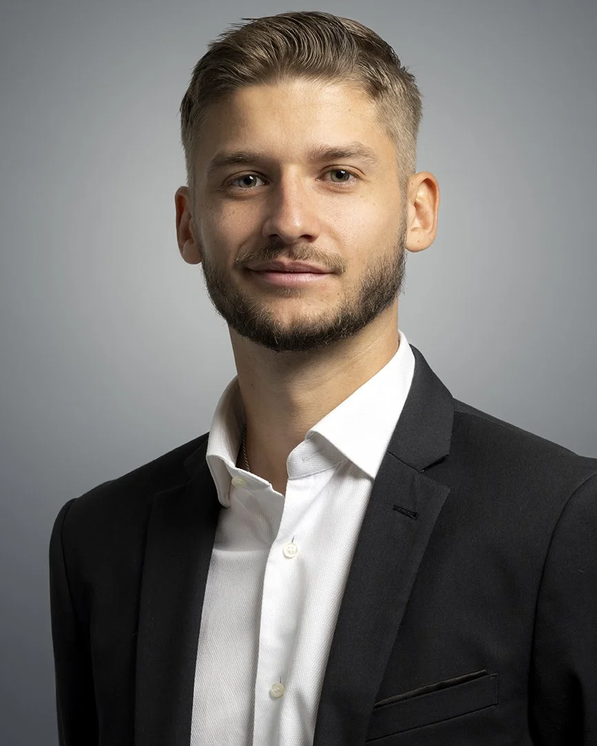 Portrait of a young man with short blonde hair, beard, wearing a black suit and white shirt, against a gray background.