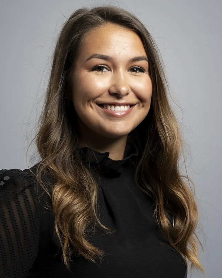 A woman with long wavy brown hair, smiling, wearing a black top with sheer details on the sleeves, against a gray background.