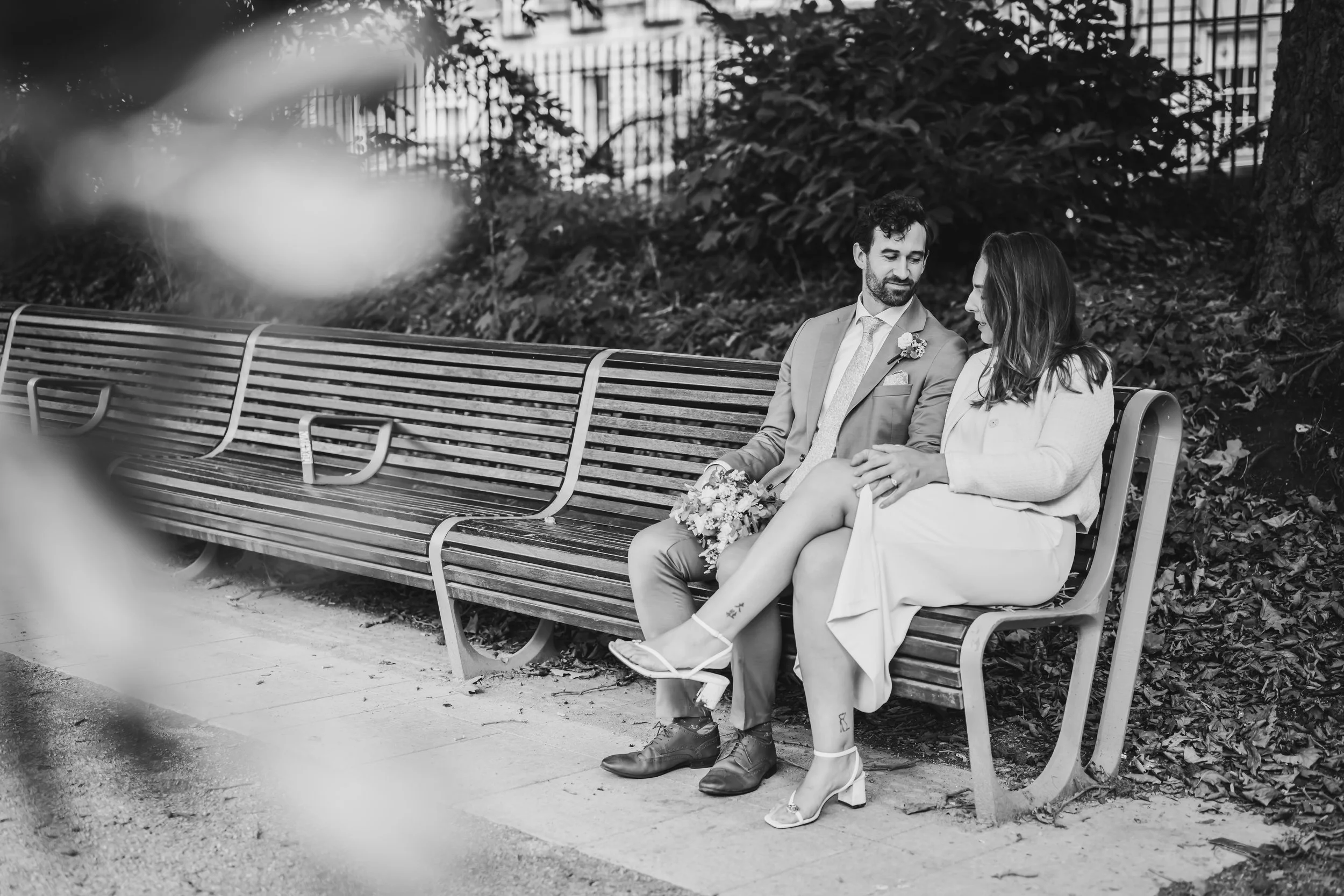 A black and white photo of a groom in a suit and a bride in a dress sitting on a park bench, engaging in conversation, with the woman holding a bouquet of flowers.